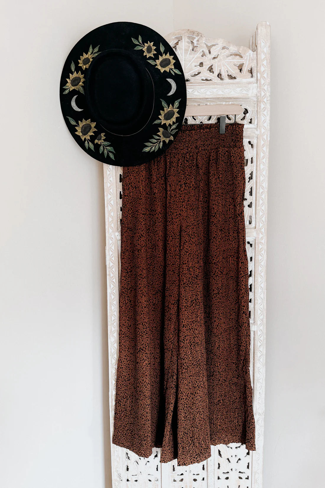 Brown high rise wide leg pants with subtle print hung on carved white wooden panel beside black hat with sunflower and moon embroidery