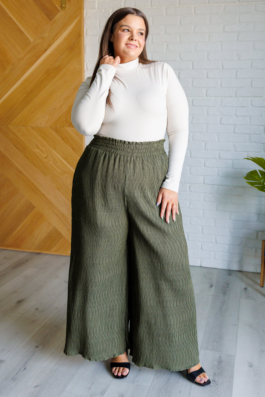 Woman wearing a white turtle neck top, olive green wide leg pants and black sandals standing against a white brick wall.