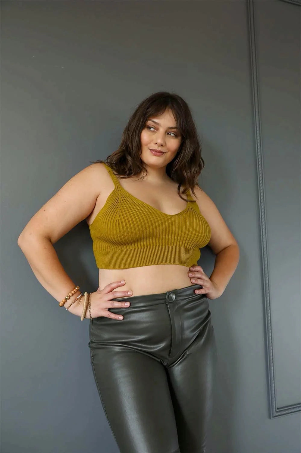 Curvy model wearing mustard hazy baby rib knit brami bralette paired with high-waisted black faux leather pants against gray wall