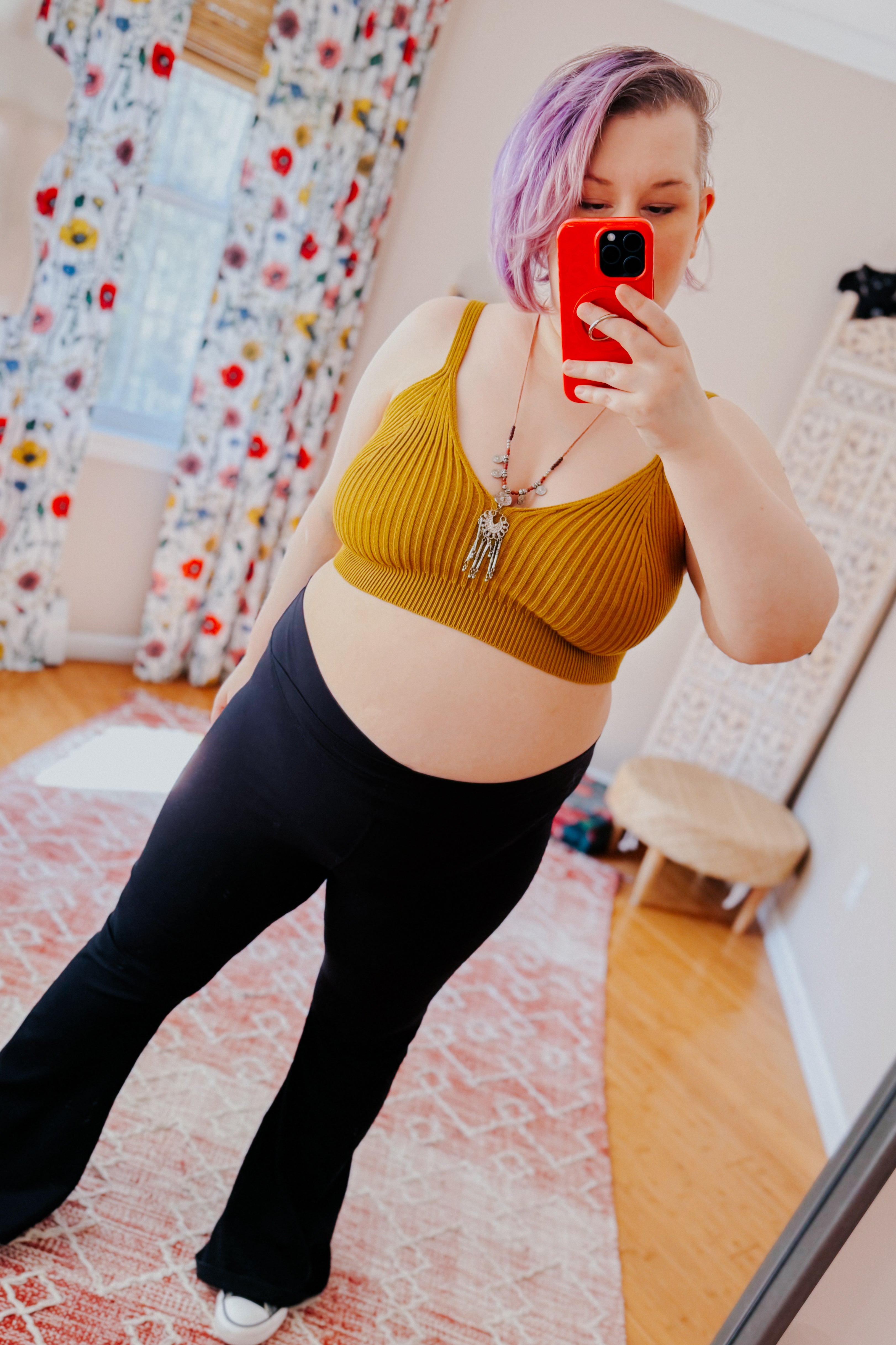 Woman with short pastel purple hair taking mirror selfie wearing mustard rib knit brami bralette and black pants indoors