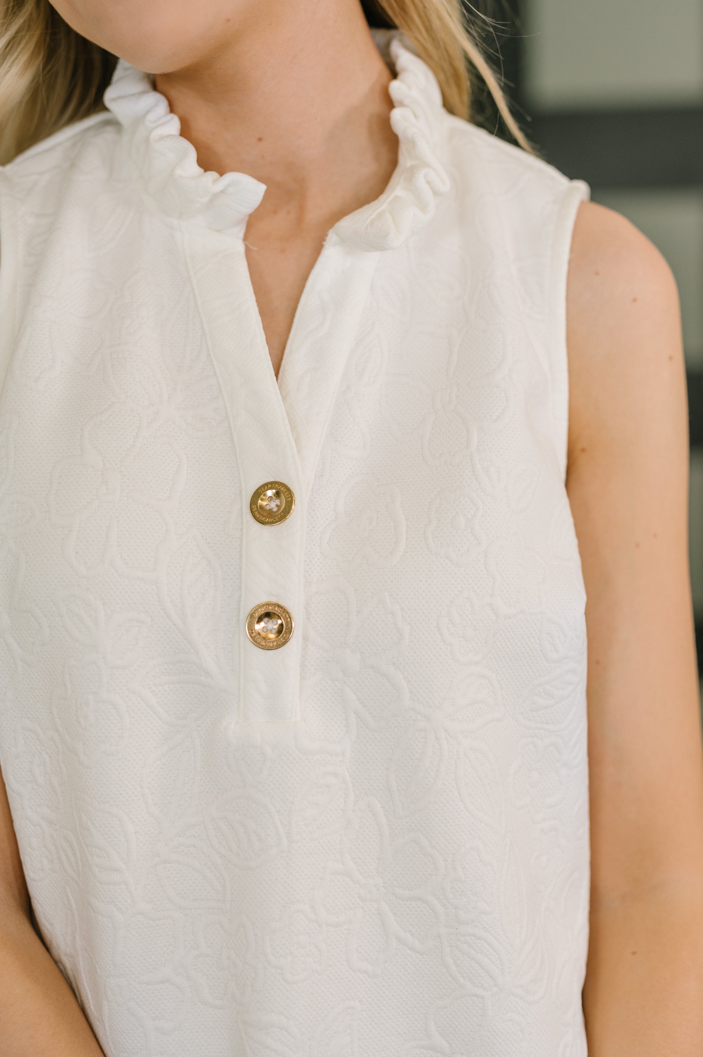 Close-up of off white sleeveless dress with ruffle high neck, gold buttons, and embossed floral knit fabric