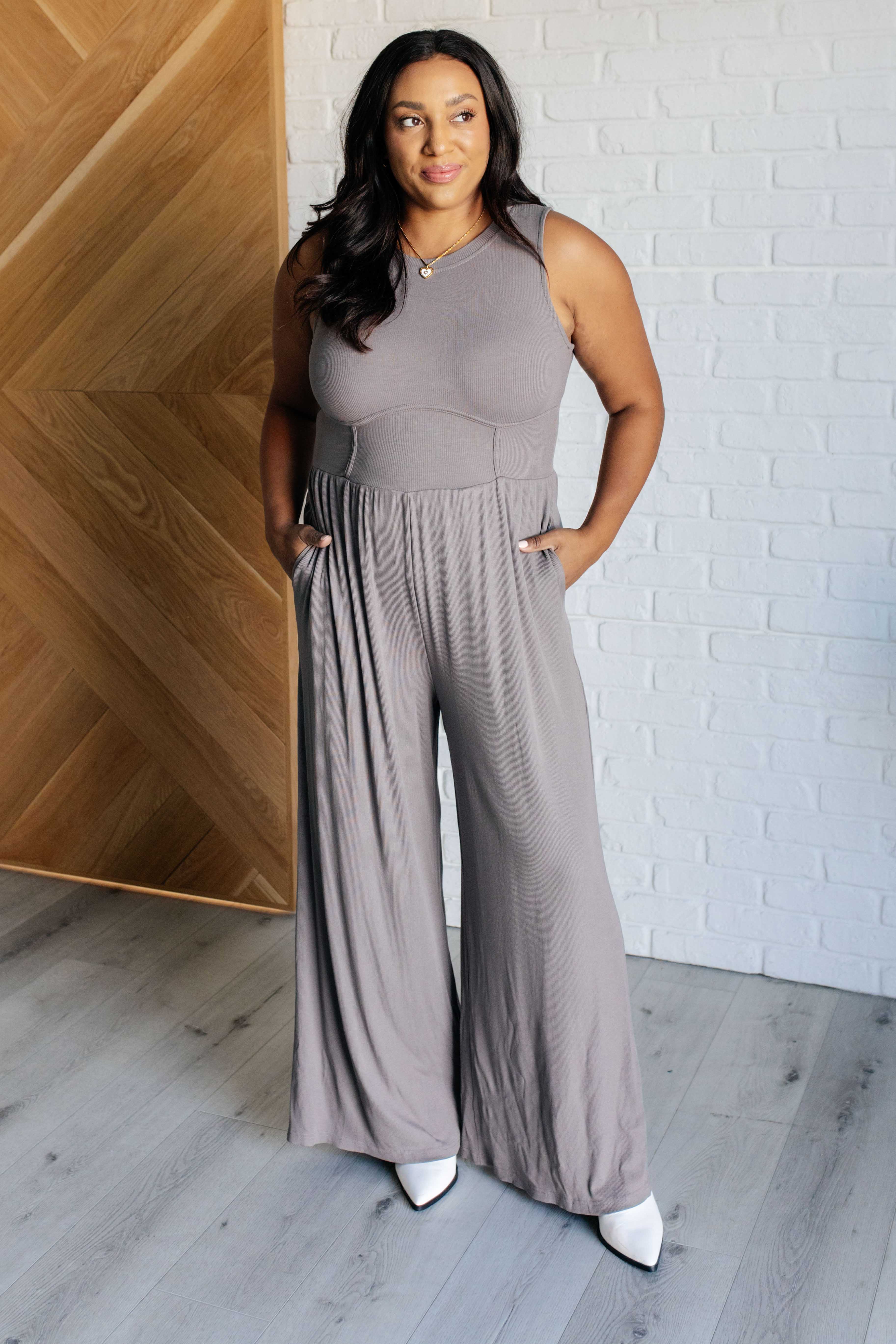 Woman wearing sleeveless grey wide leg jumpsuit with fitted waist and pockets standing indoors by white brick wall and wooden door