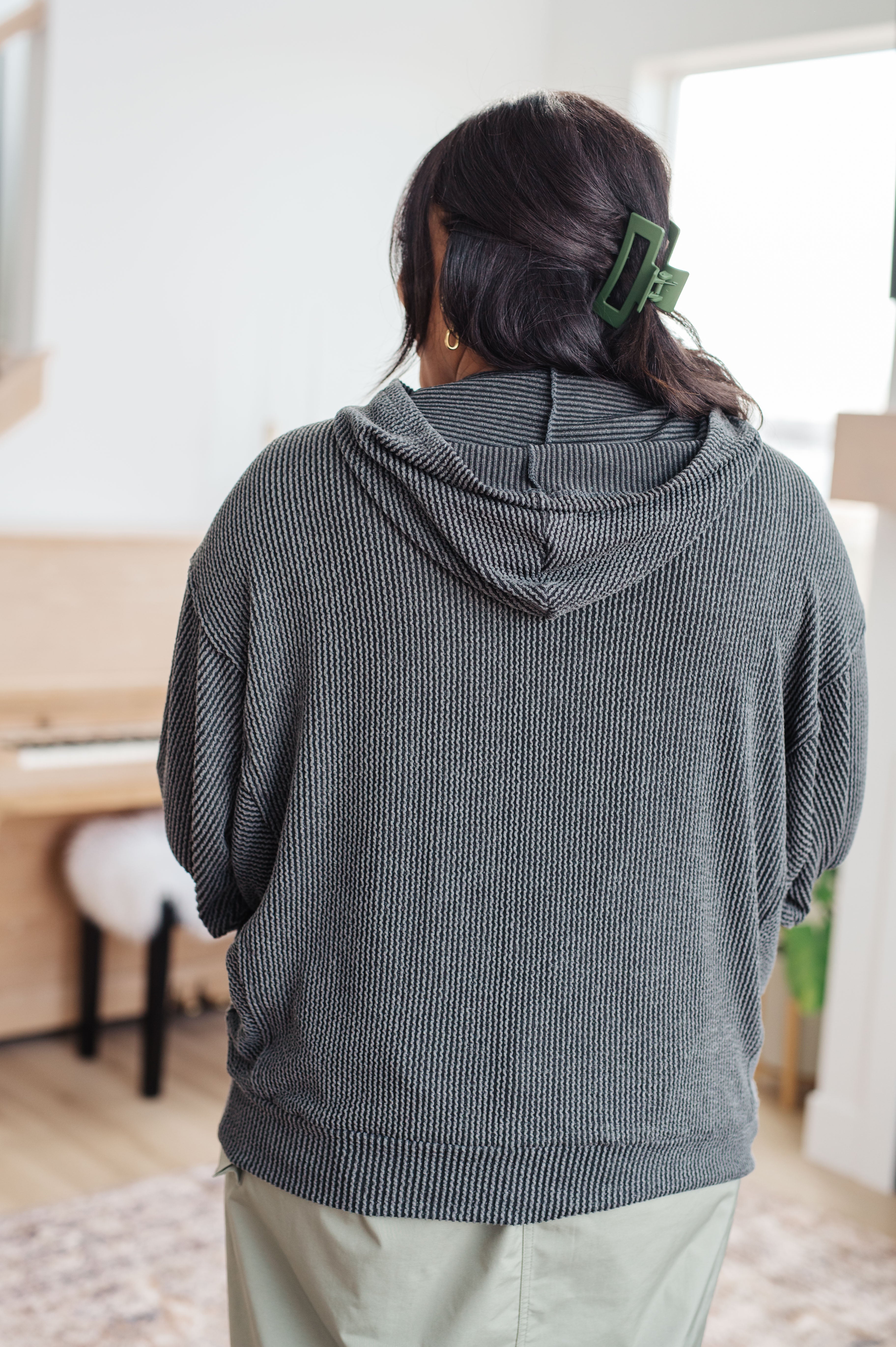 Woman with her back facing the camera wearing a grey rib knit hoodie over a green skirt against an indoor setting.