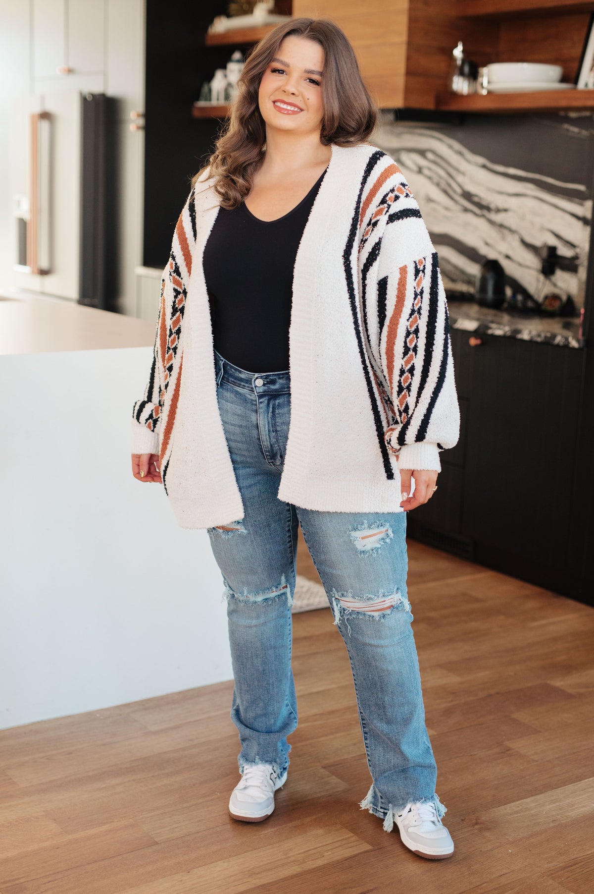 Woman wearing a cream open front knit cardigan with bold patterned sleeves styled over a black top with distressed jeans and sneakers indoors