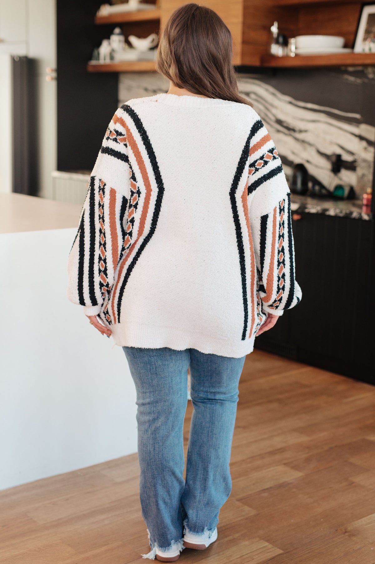 Back view of a woman wearing a cream fuzzy knit cardigan with bold black and rust patterns, blue jeans, and sneakers indoors