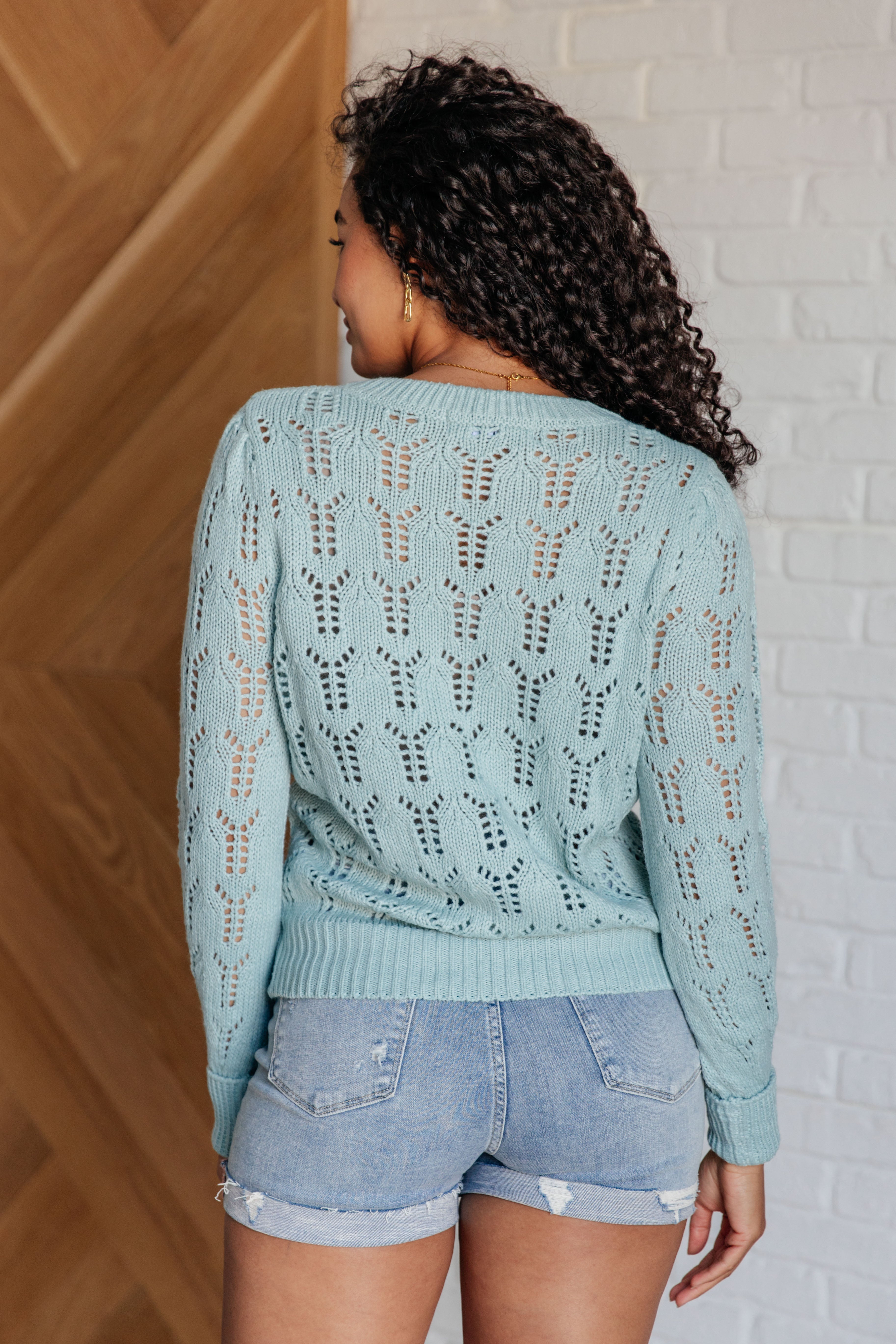 Rear view of a light blue sheer pointelle knit sweater worn with distressed denim shorts on model