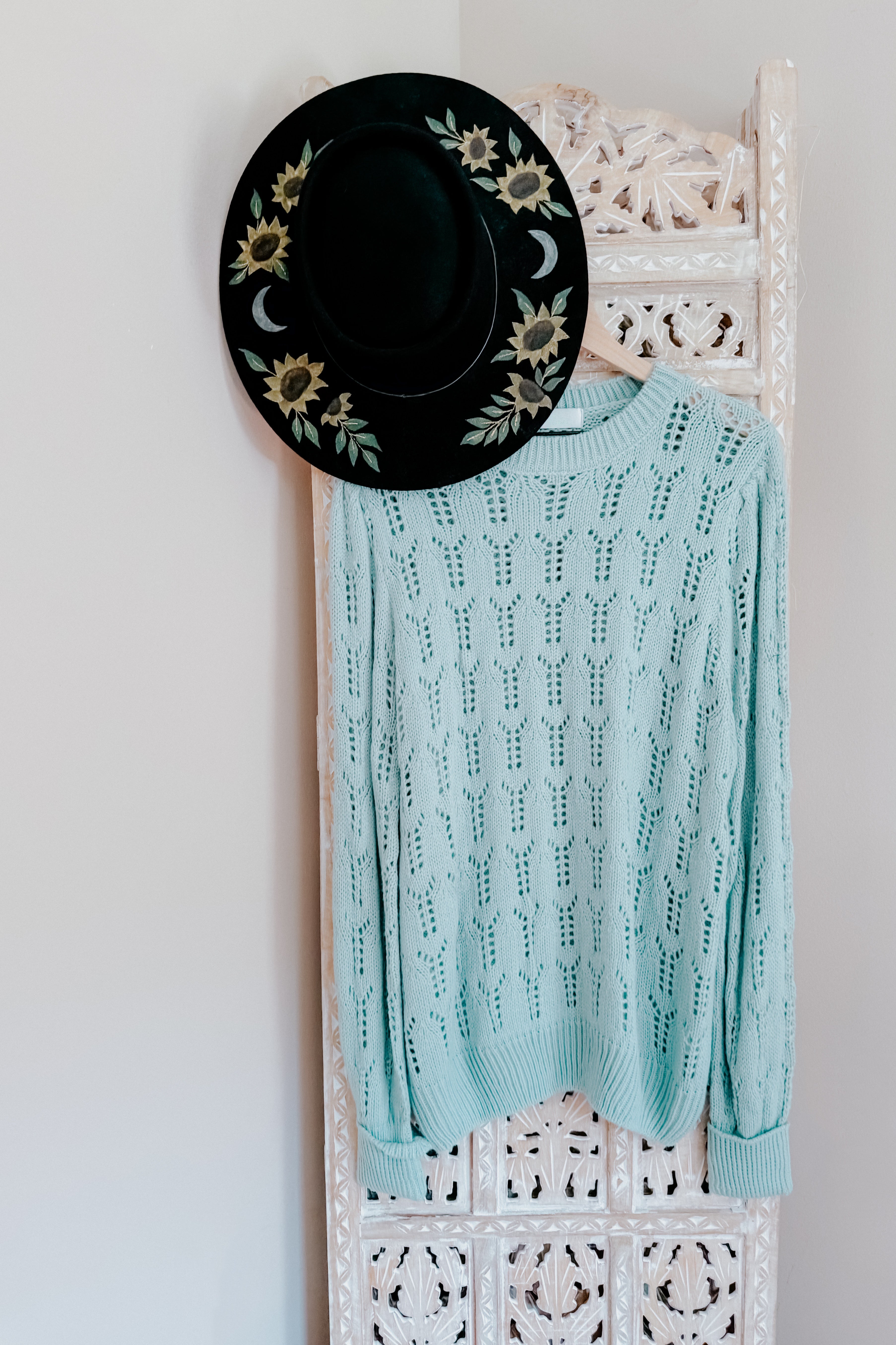 Light blue sheer pointelle knit sweater hanging on carved white screen with black floral embroidered hat above