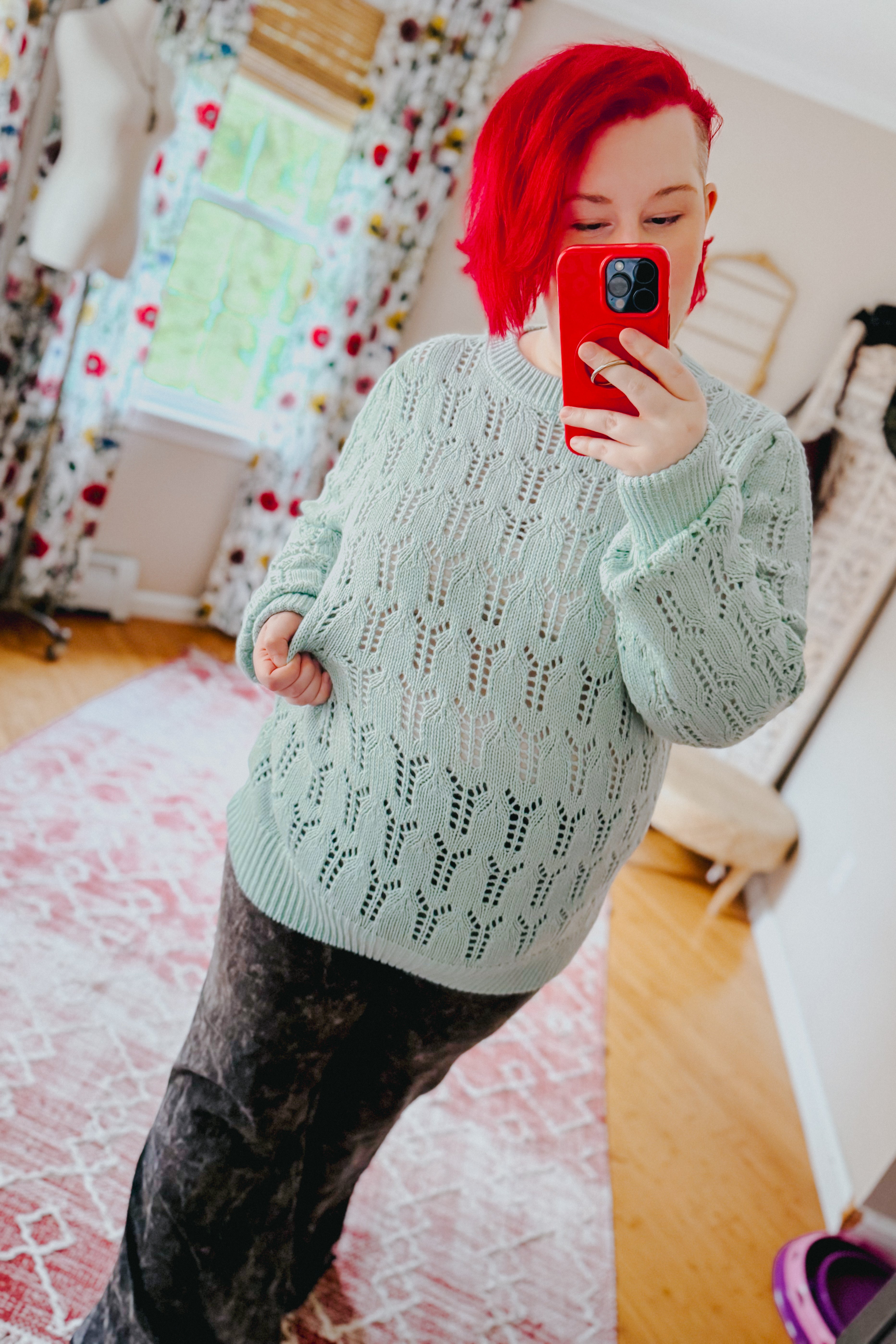 Woman with bright red hair taking mirror selfie wearing light green sheer pointelle knit sweater with long sleeves indoors