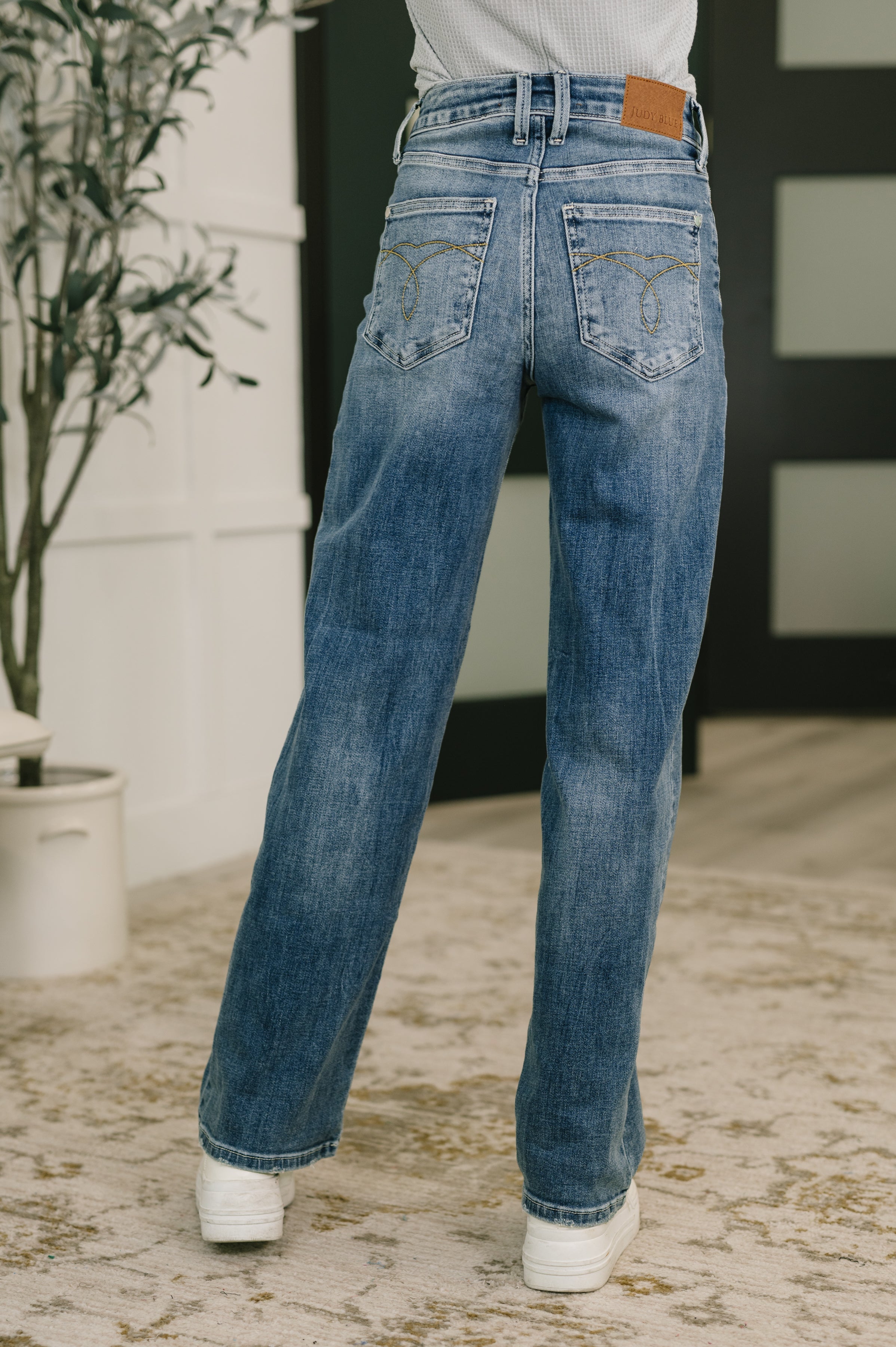 Back view of woman wearing blue mid rise straight jeans with slight fading and white sneakers indoors