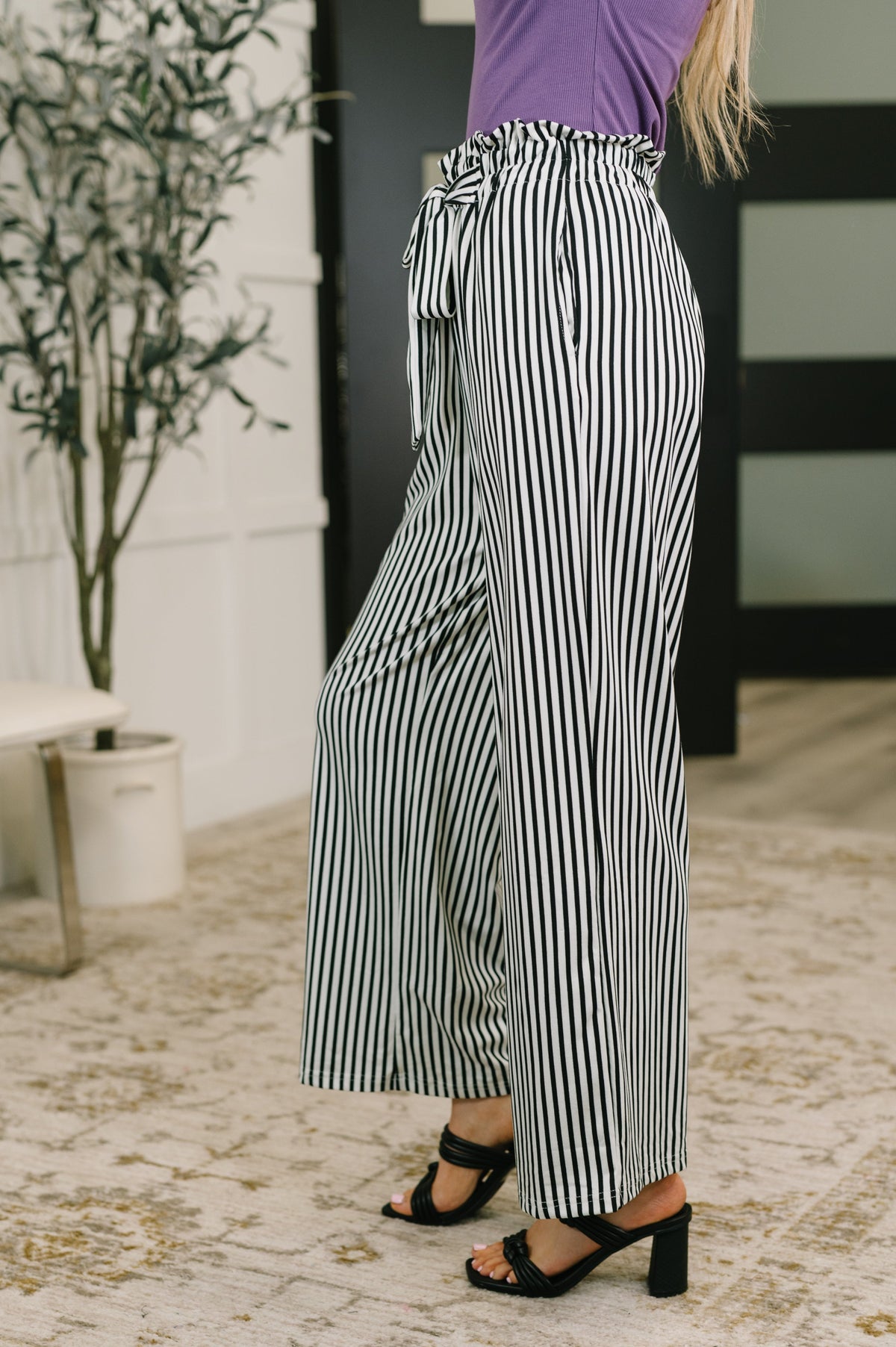 Side view of a woman wearing black and white striped high waisted wide leg trousers with a tie waist purple top and black heeled sandals indoors