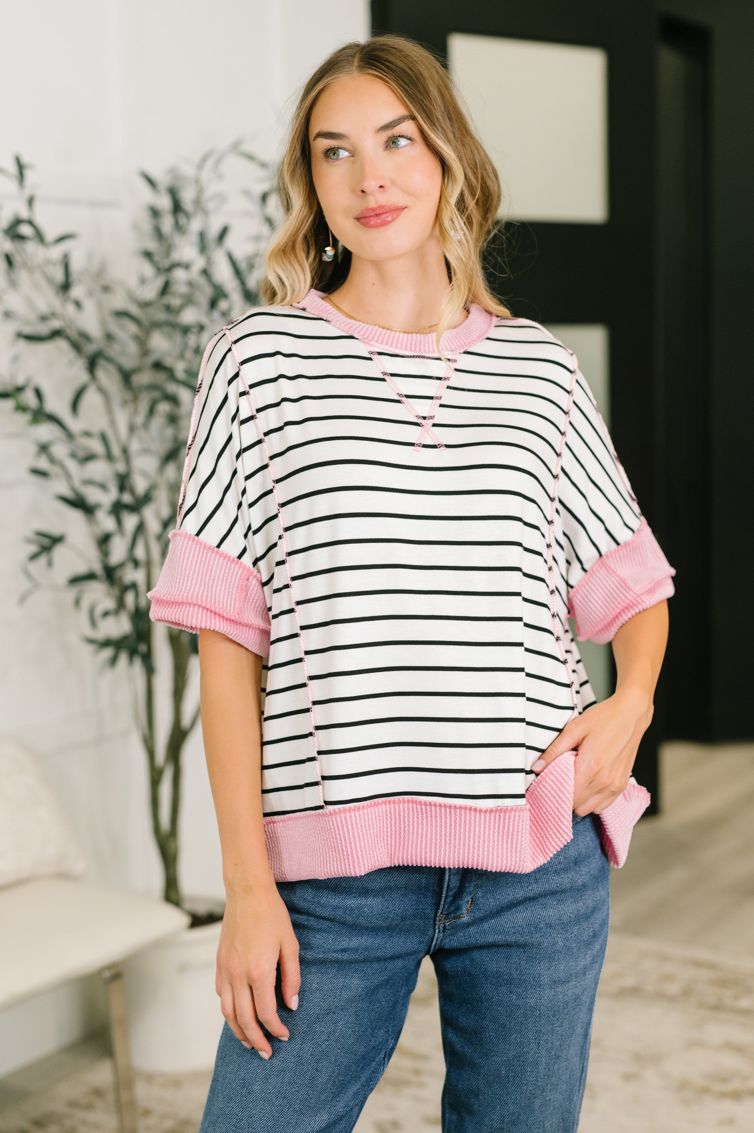 Woman wearing ivory striped boxy knit top with pink contrast ribbed cuffs and hem paired with blue jeans indoors
