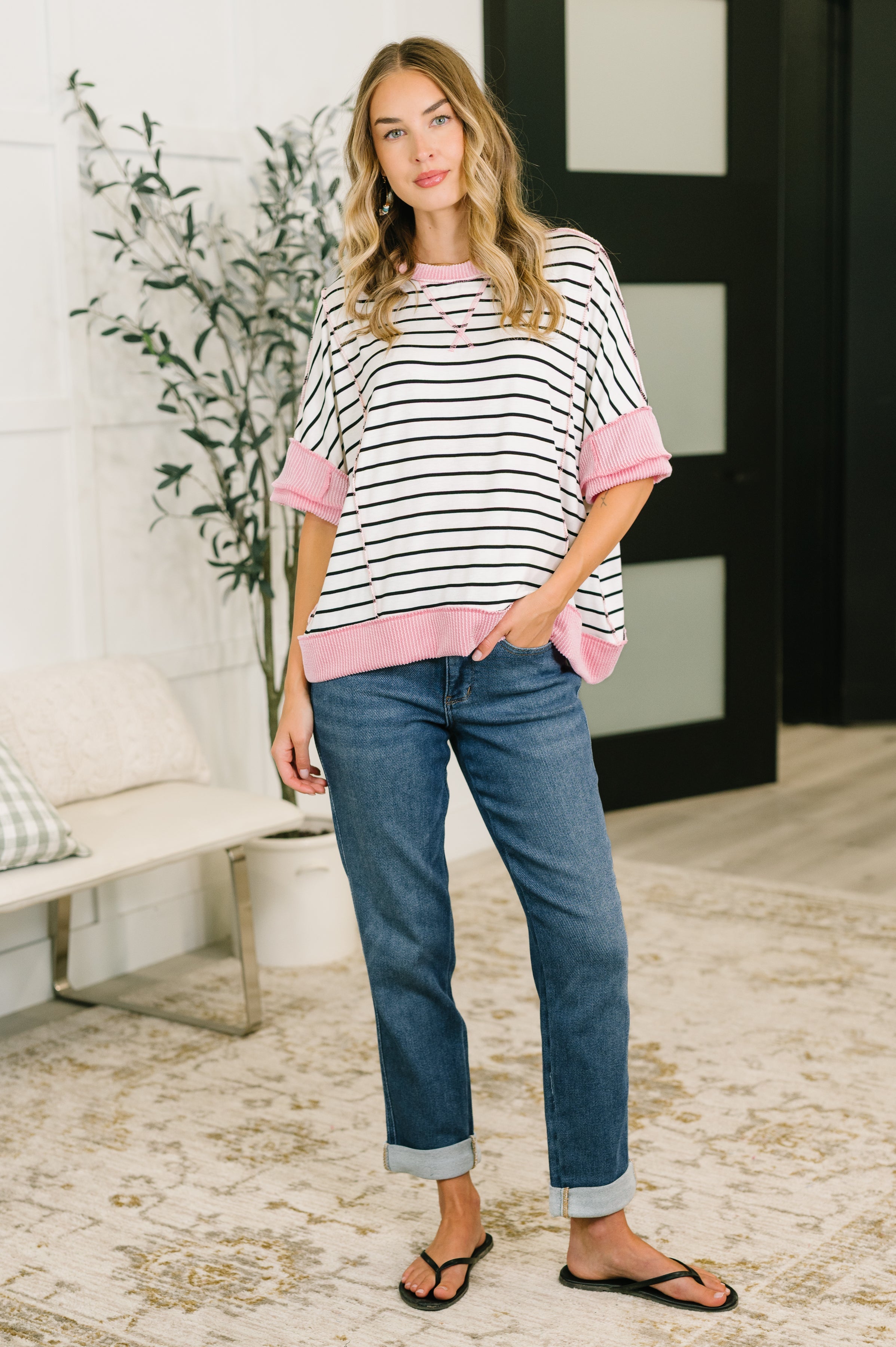 Woman wearing ivory striped top with pink ribbed trim and cuffed blue jeans standing indoors with hand in pocket