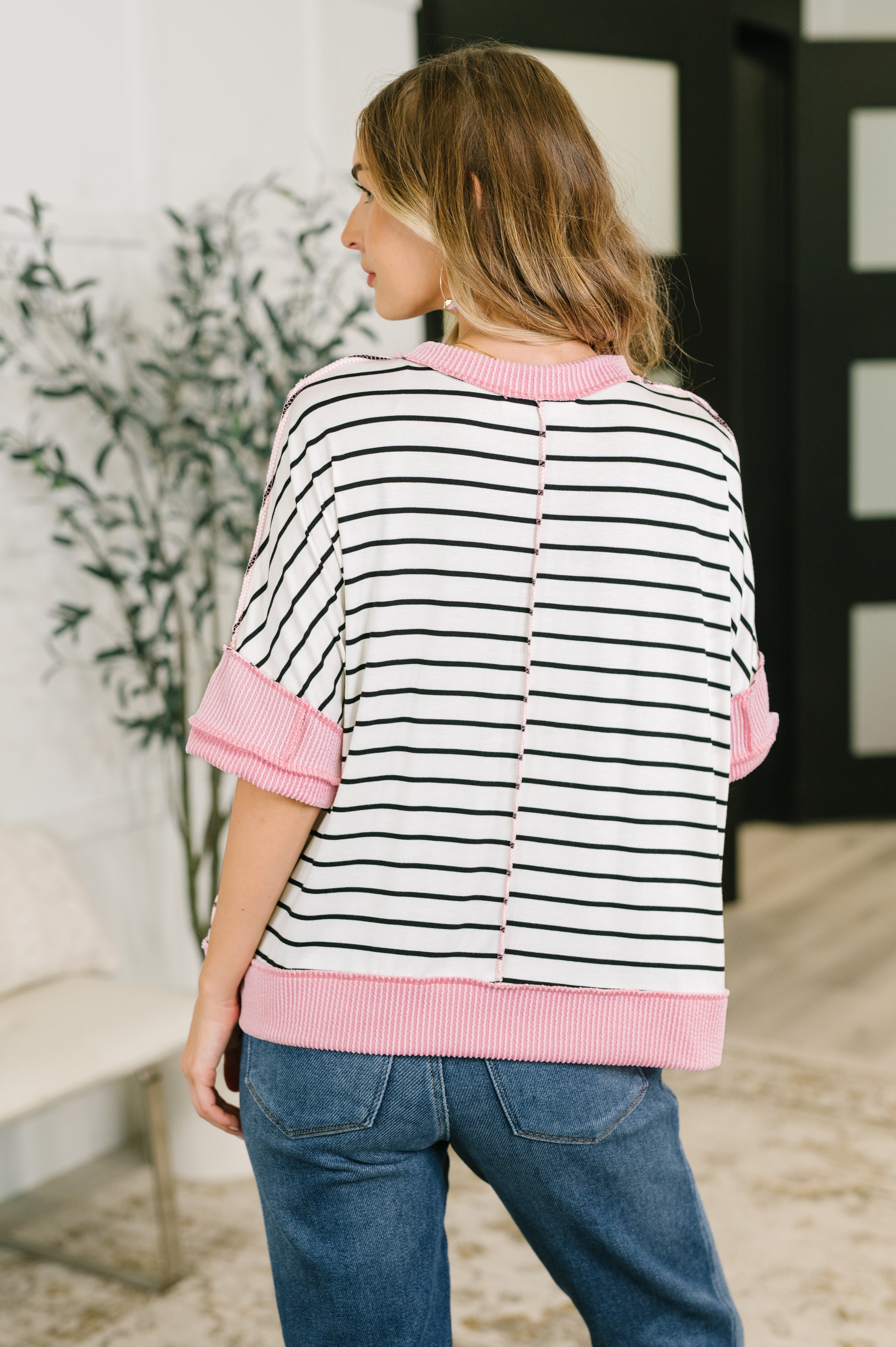 Woman wearing ivory striped boxy top with pink ribbed collar, cuffs, and hem paired with blue jeans indoors, back view