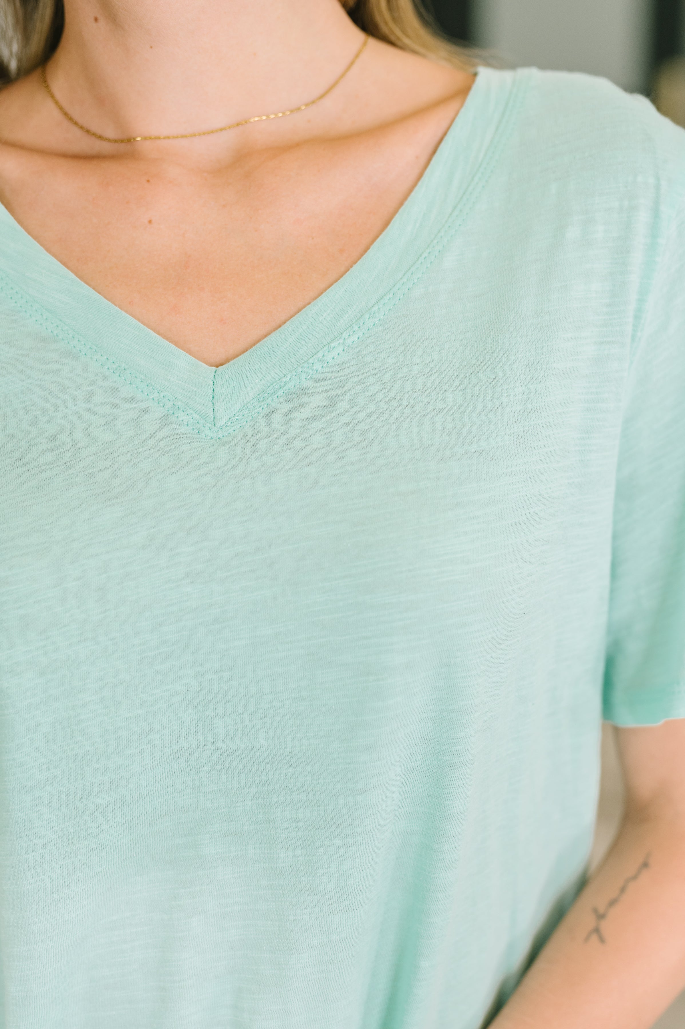 Close-up of soft heathered mint knit V-neck tee with relaxed fit and delicate gold chain necklace worn indoors