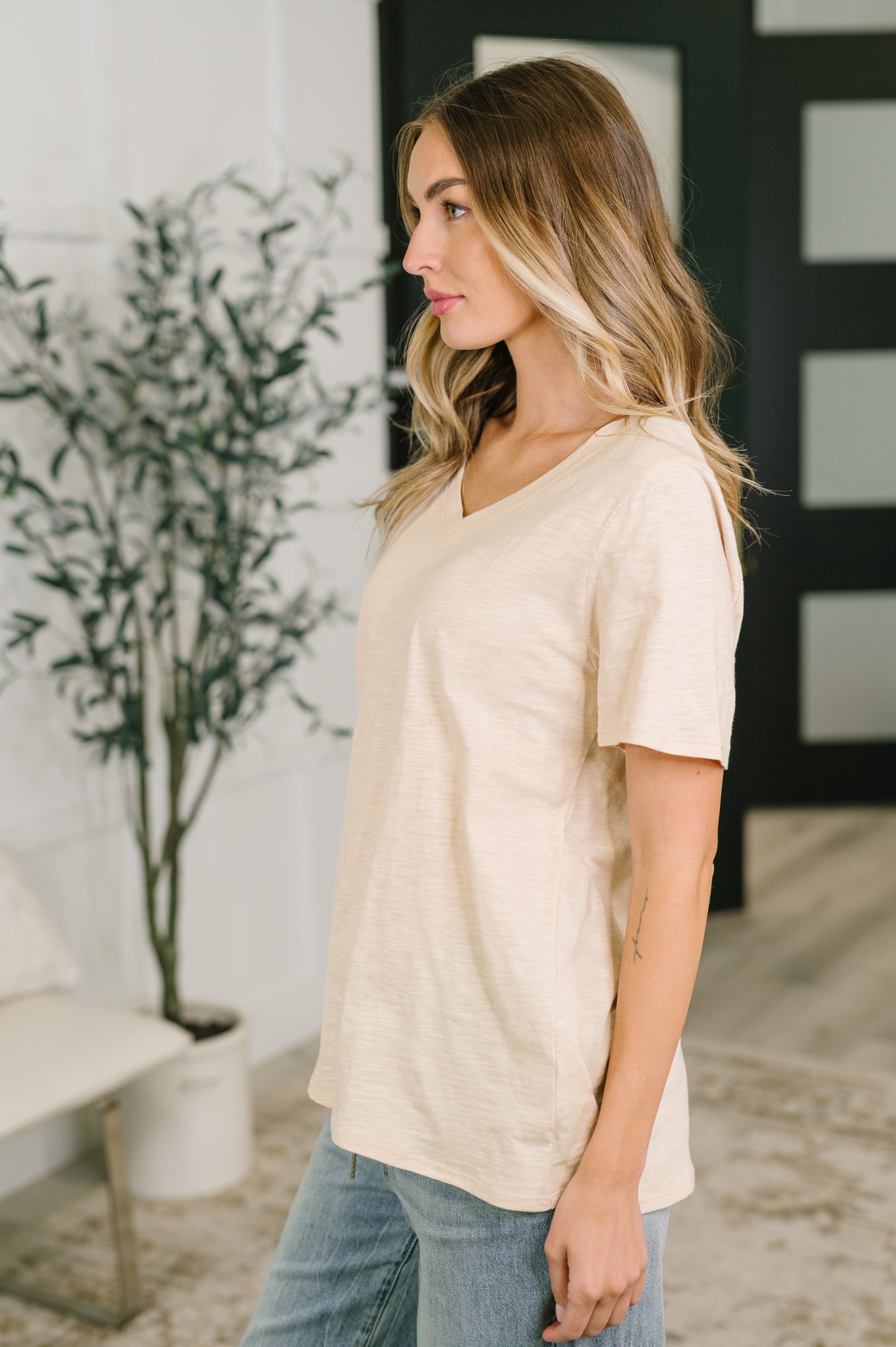 Woman wearing oatmeal V-neck tee with relaxed fit paired with light blue jeans indoors by plant and bench