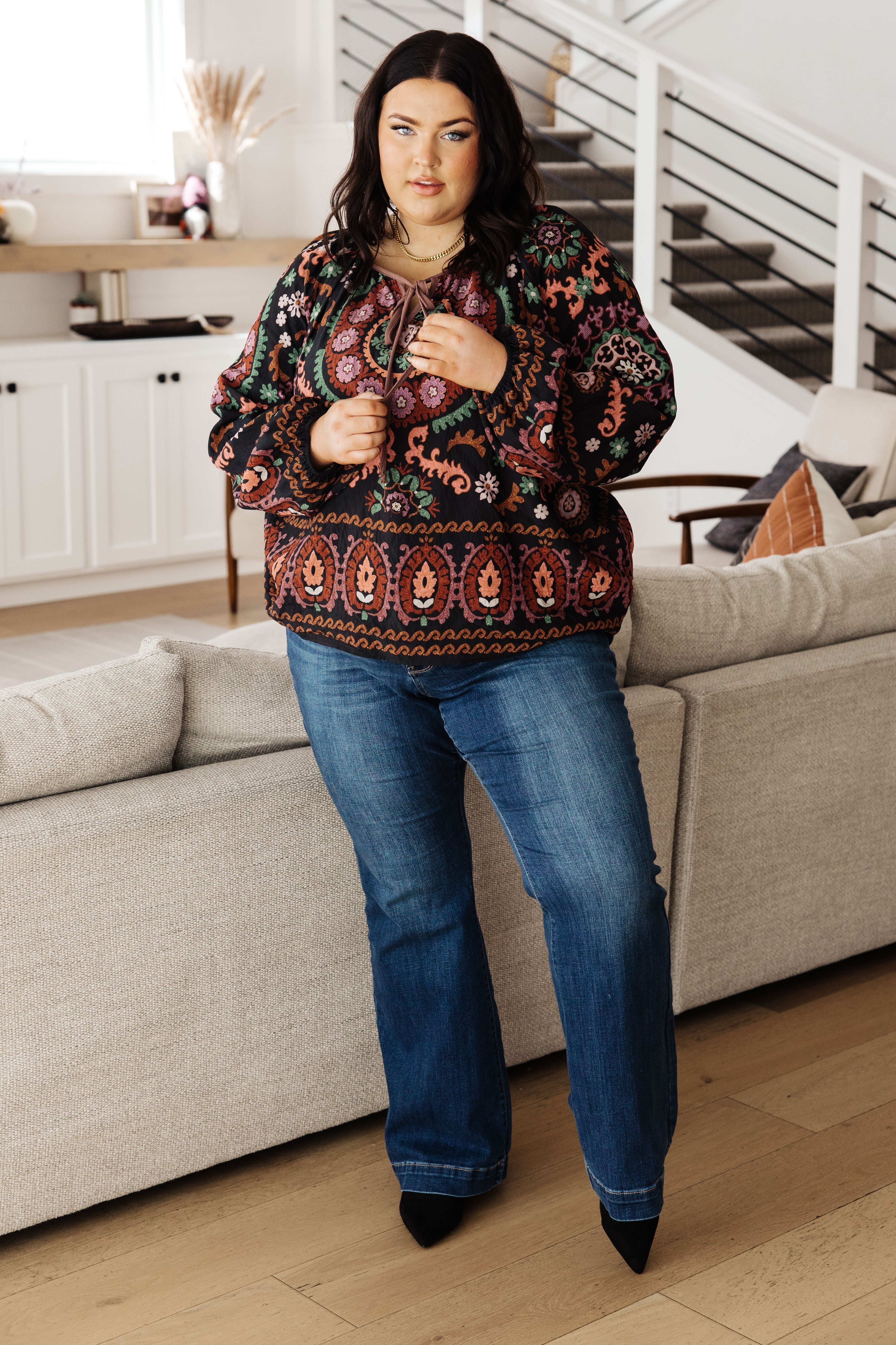 Woman wearing a black I Feel That Peasant Blouse with colorful patterns paired with blue denim jeans indoors by beige couch
