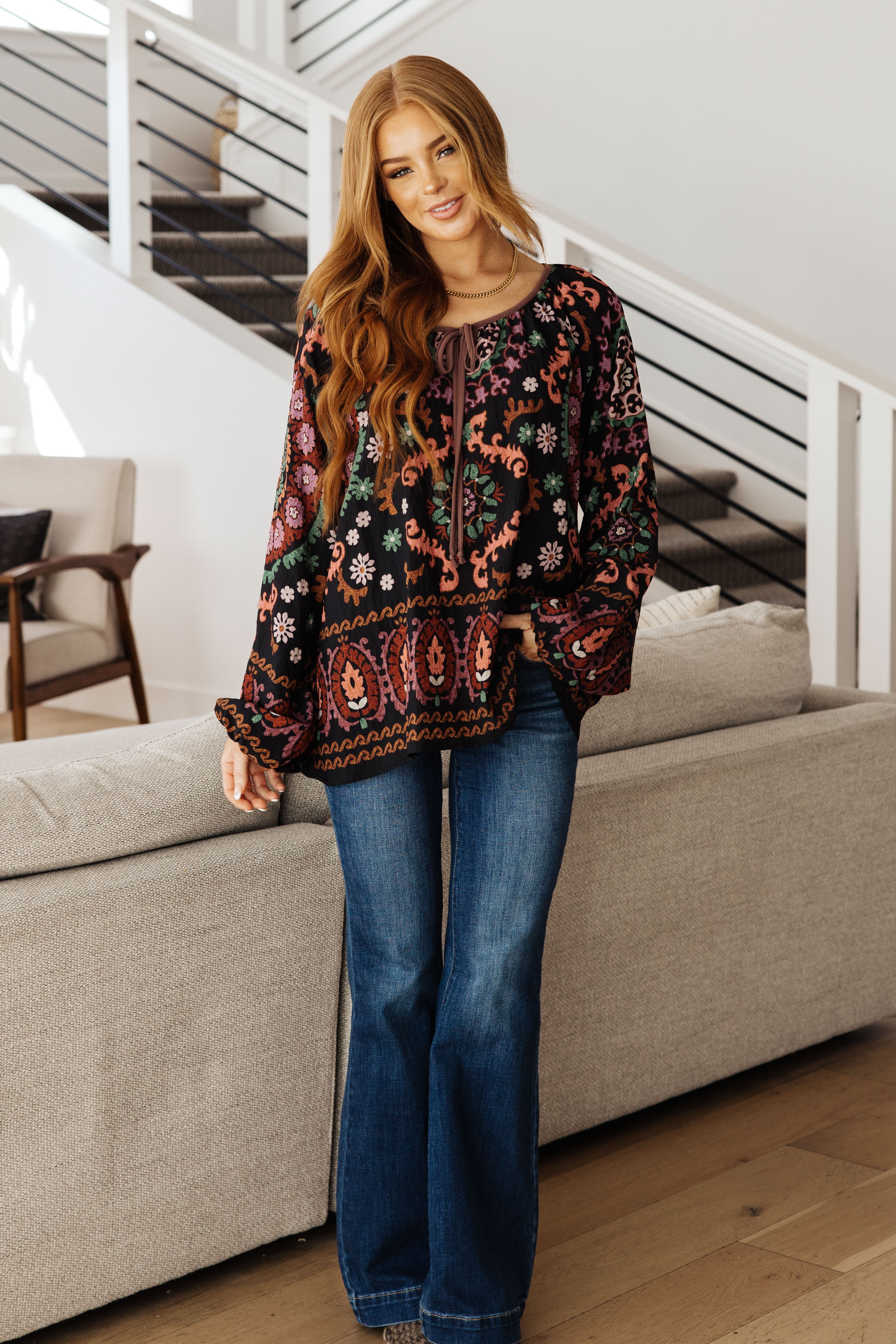 Woman standing indoors wearing black I Feel That Peasant Blouse with detailed colorful paisley and floral designs paired with blue jeans