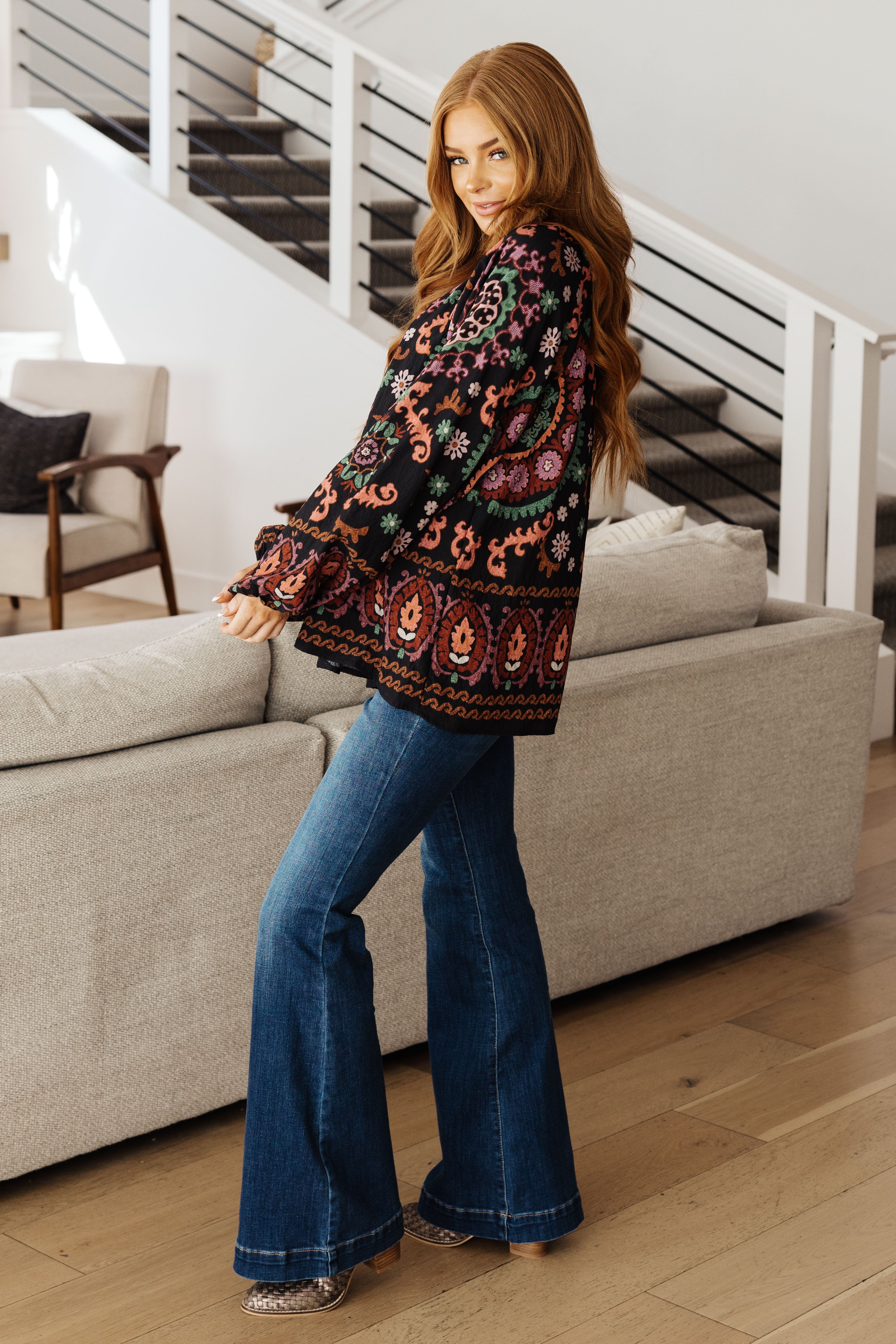 Woman indoors wearing black I Feel That Peasant Blouse with vibrant paisley patterns paired with blue flared jeans and boots