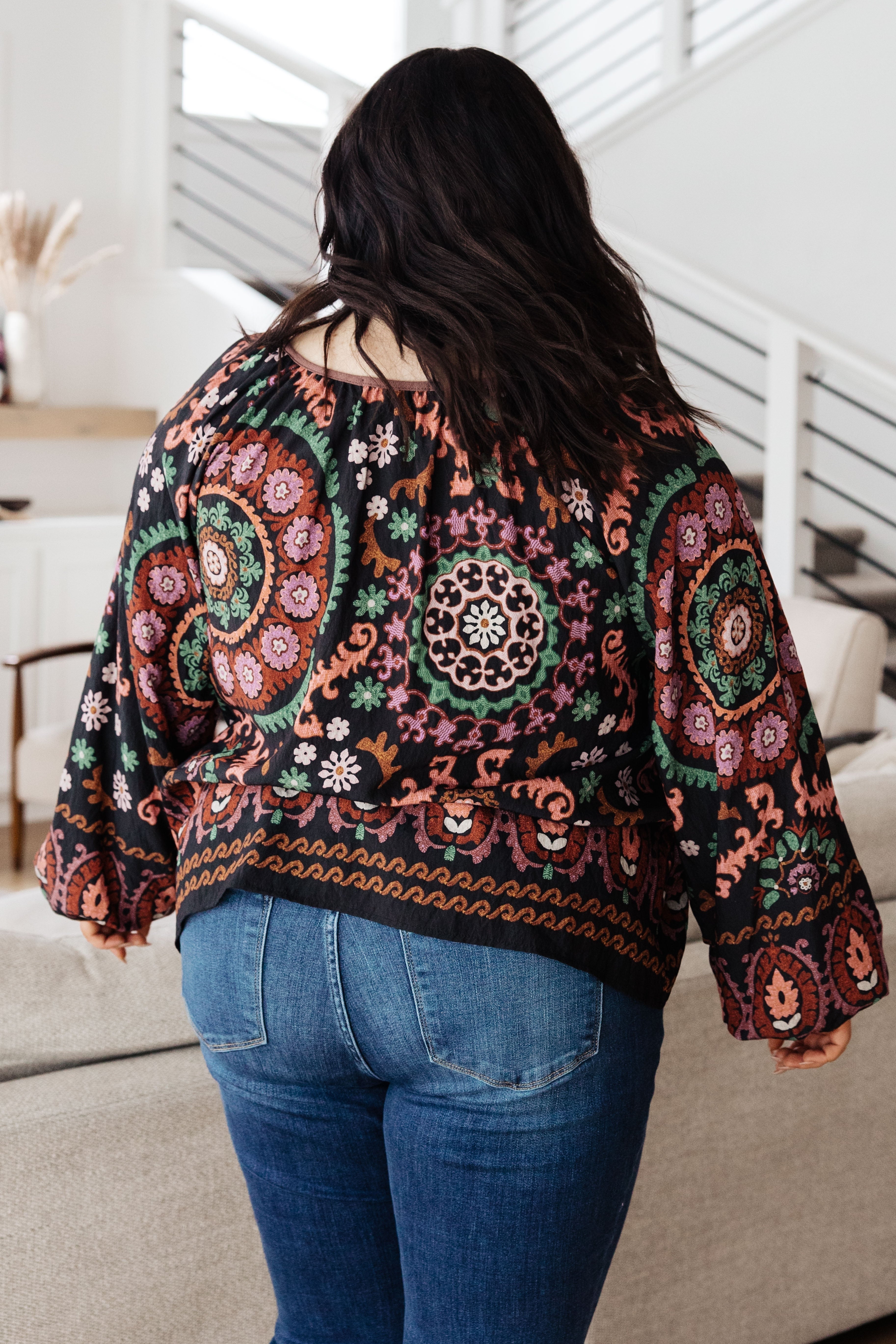 Back view of woman wearing black I Feel That Peasant Blouse with intricate colorful circular patterns paired with blue jeans indoors