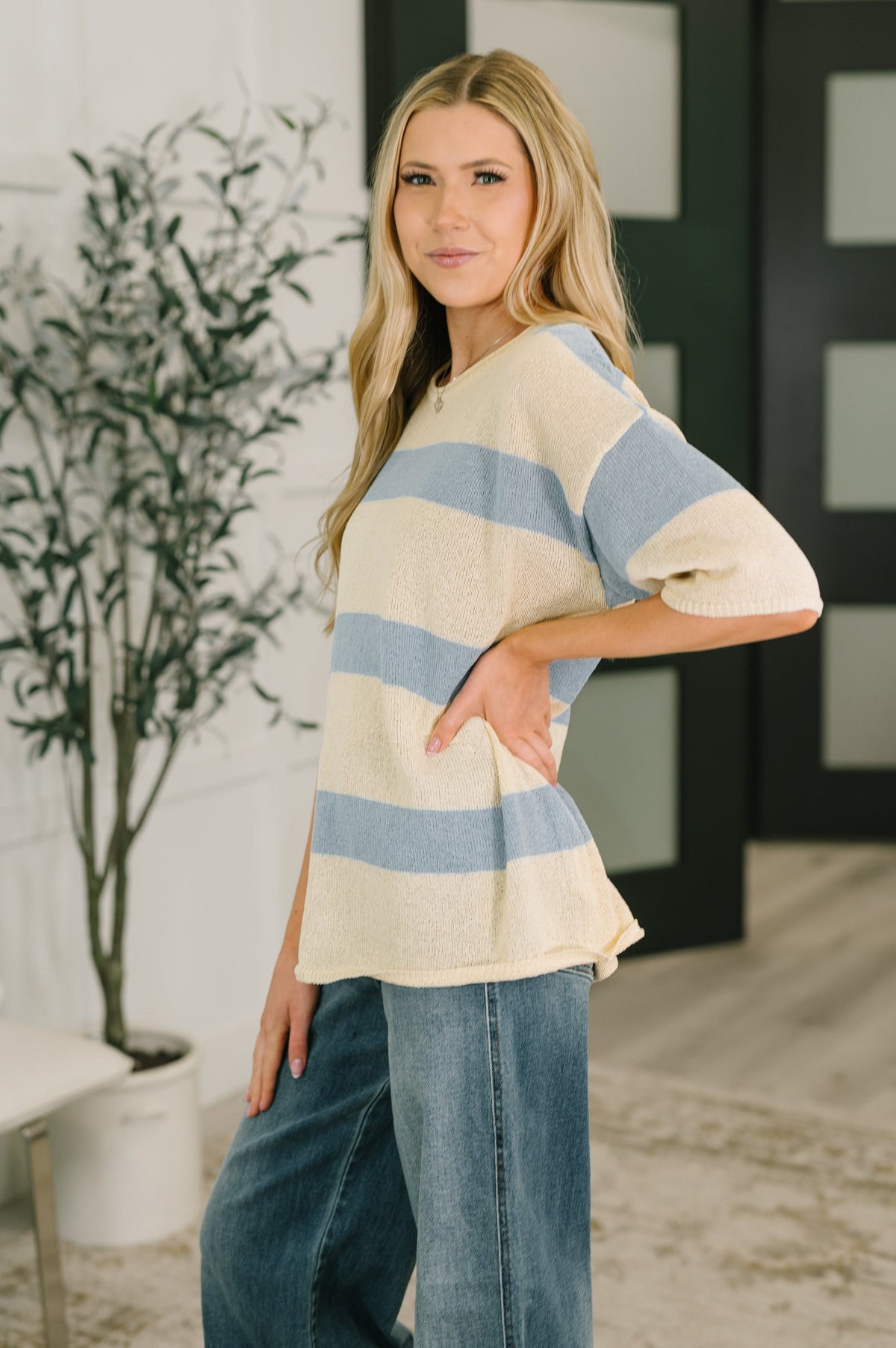 Woman modeling a beige lightweight knit top with blue horizontal stripes and relaxed drop shoulders over wide leg jeans indoors