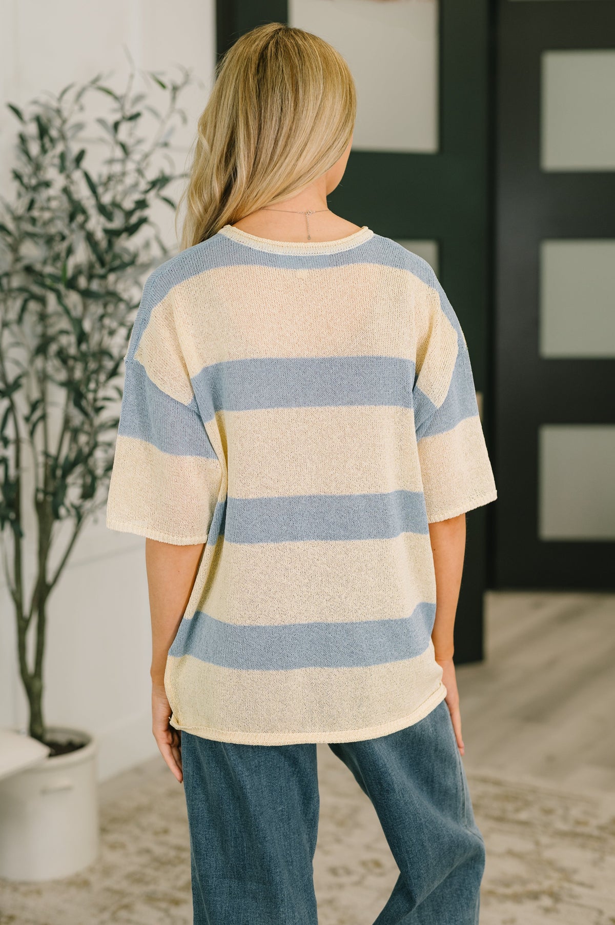 Back view of a woman wearing a beige and blue striped lightweight knit drop shoulder top with relaxed sleeves and blue jeans indoors