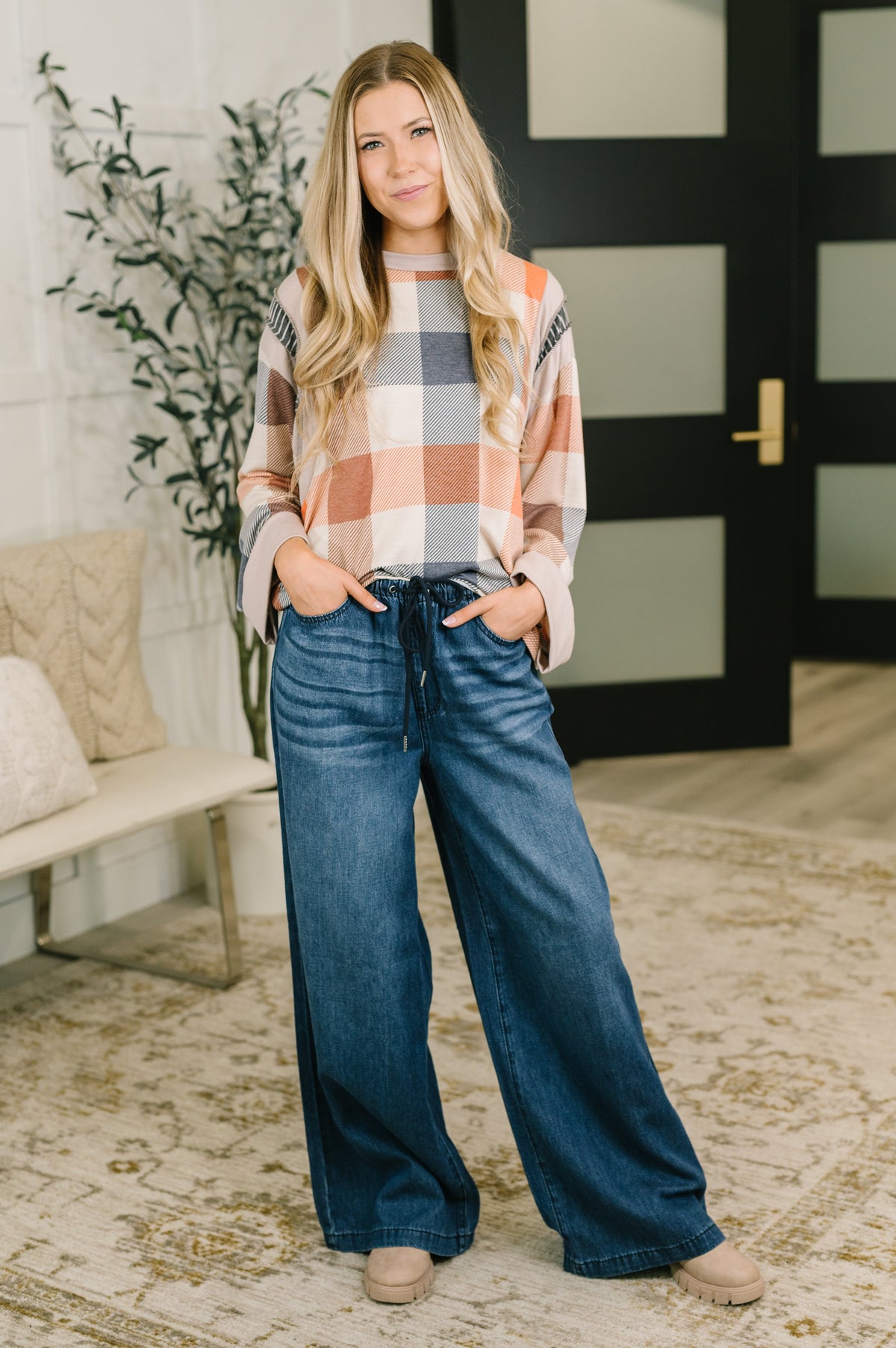 Woman in an orange and multicolor plaid long sleeve top poses with wide leg blue jeans beside a modern black door