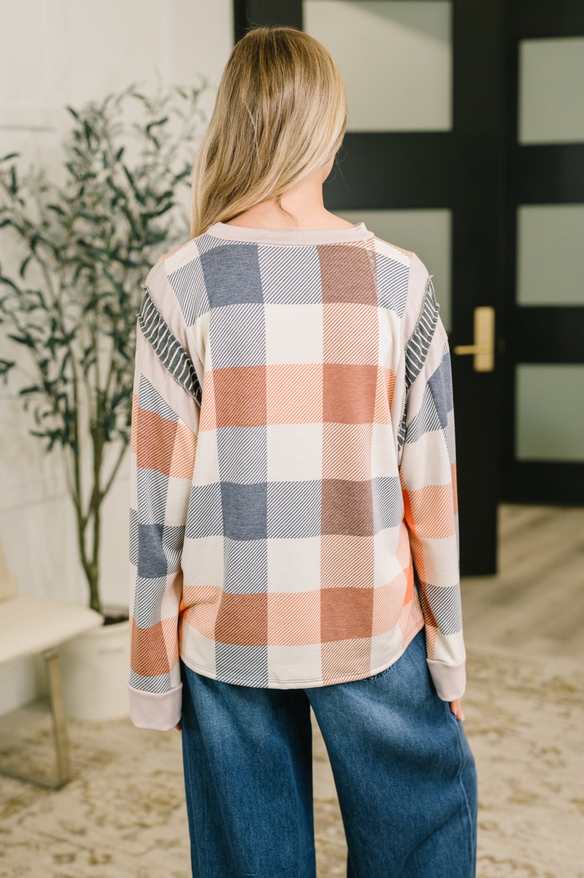 Back view of a woman wearing an orange plaid relaxed top with long sleeves and wide leg blue jeans in a stylish entryway