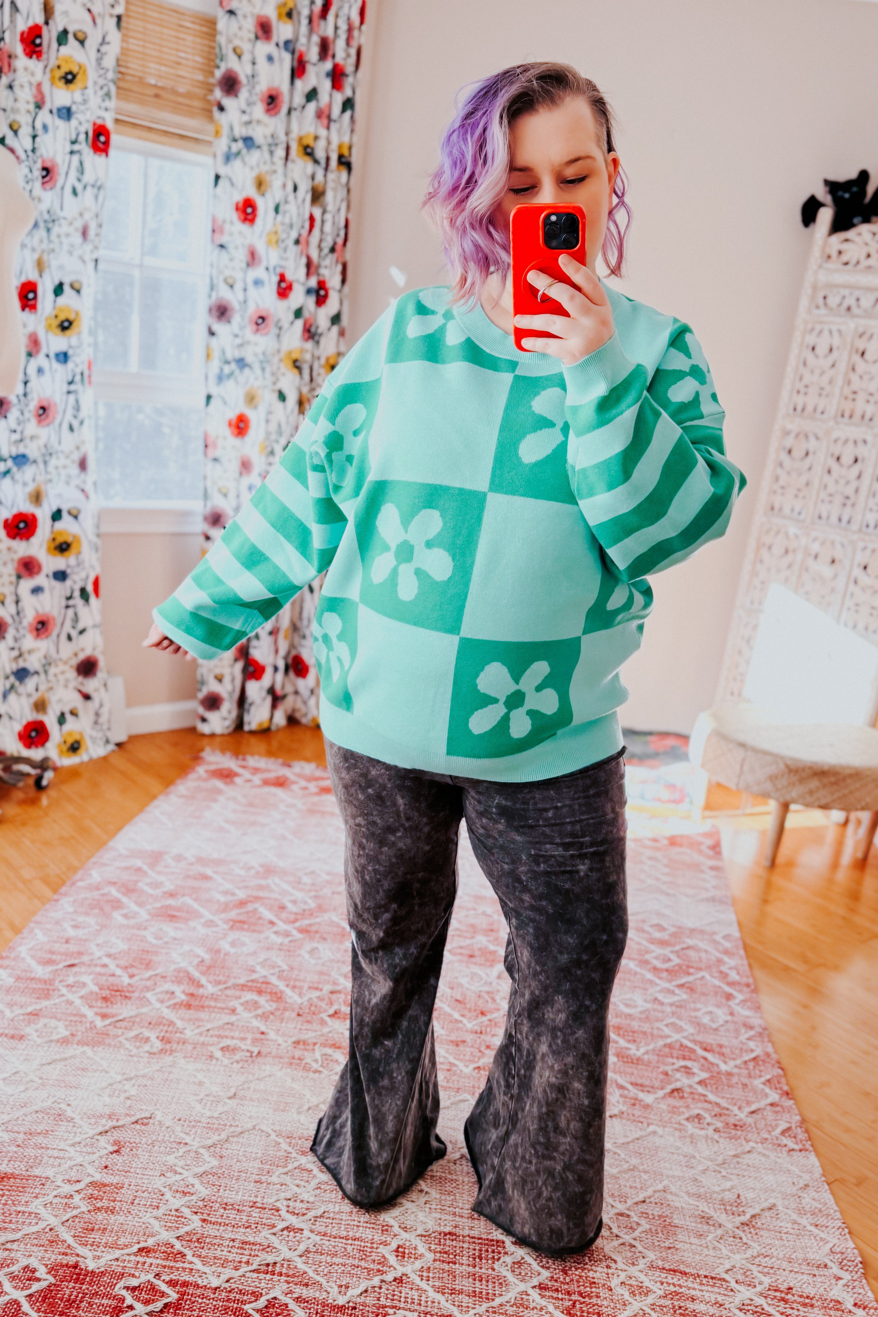 Woman wearing a checkered floral print mint sweater over acid wash flared pants in an indoor setting.