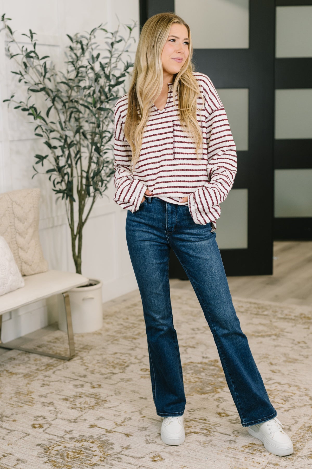 Woman wearing a cream and red striped textured knit long sleeve top with jeans and white sneakers indoors