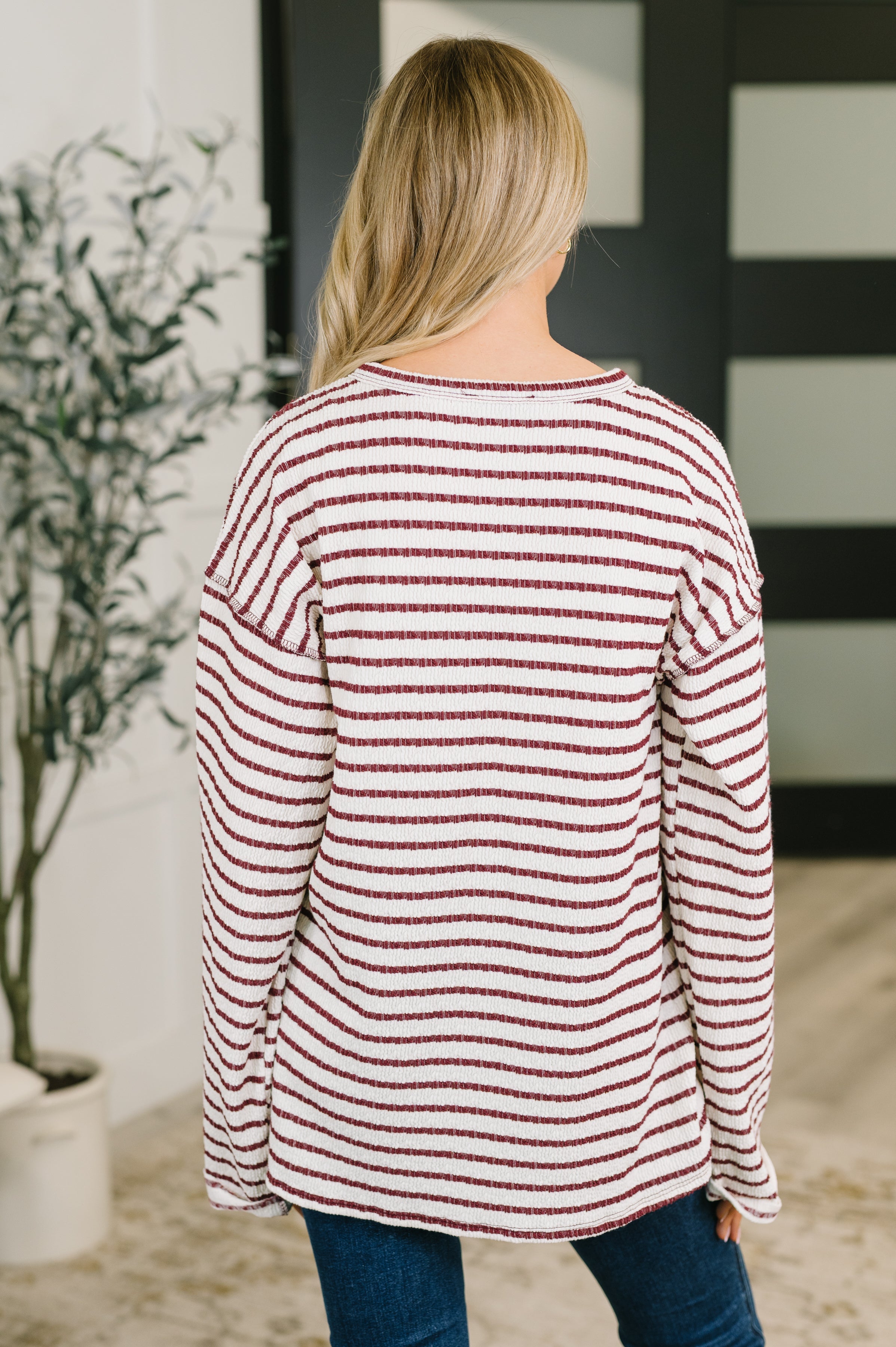 Back view of a woman wearing a cream textured knit long sleeve top with burgundy horizontal stripes and jeans indoors