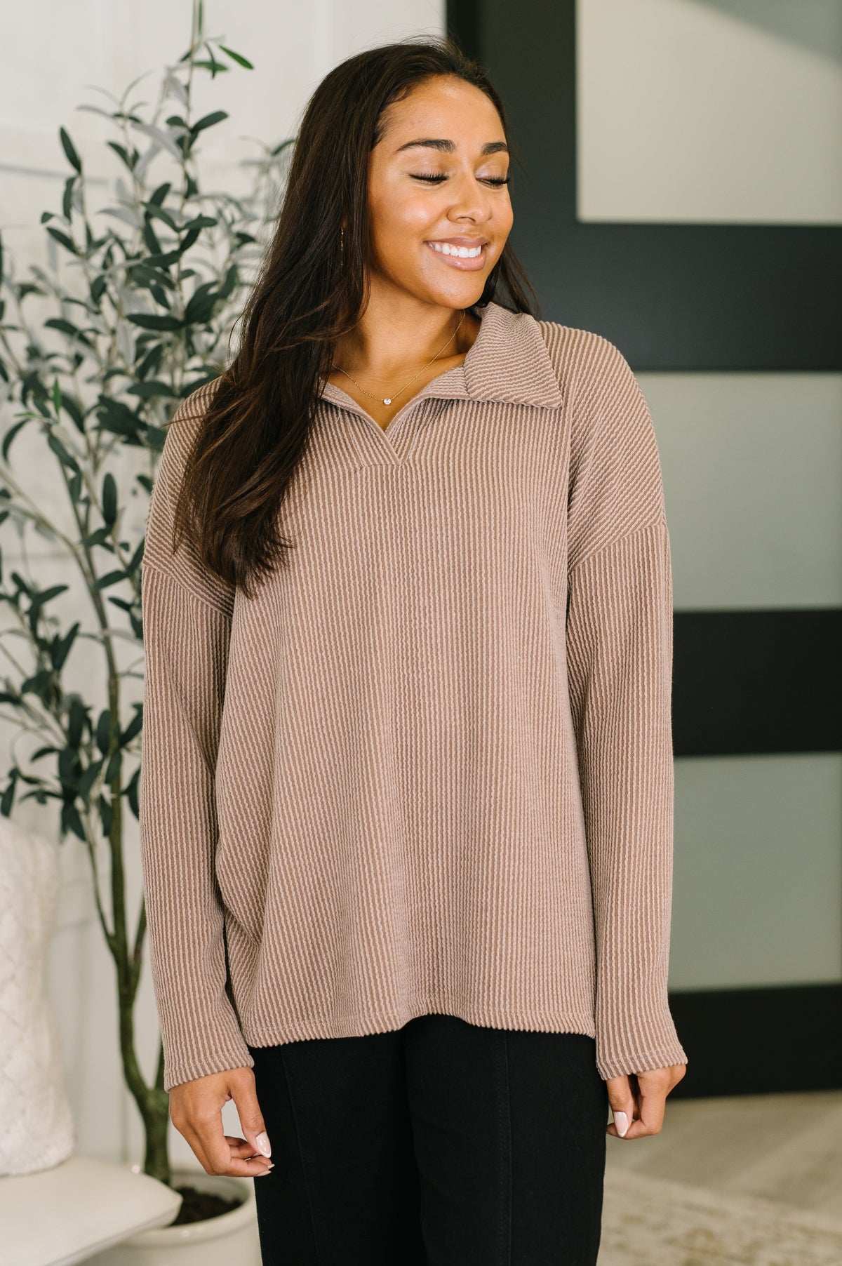 Smiling woman modeling a mocha textured ribbed collared pullover with relaxed sleeves and black pants in a modern home interior