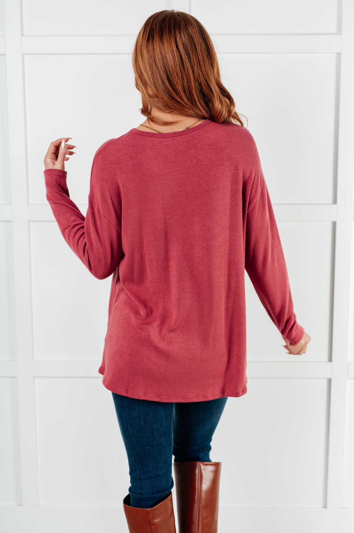 Back view of a woman in a cranberry drop shoulder long sleeve top with dark jeans and brown knee high boots indoors