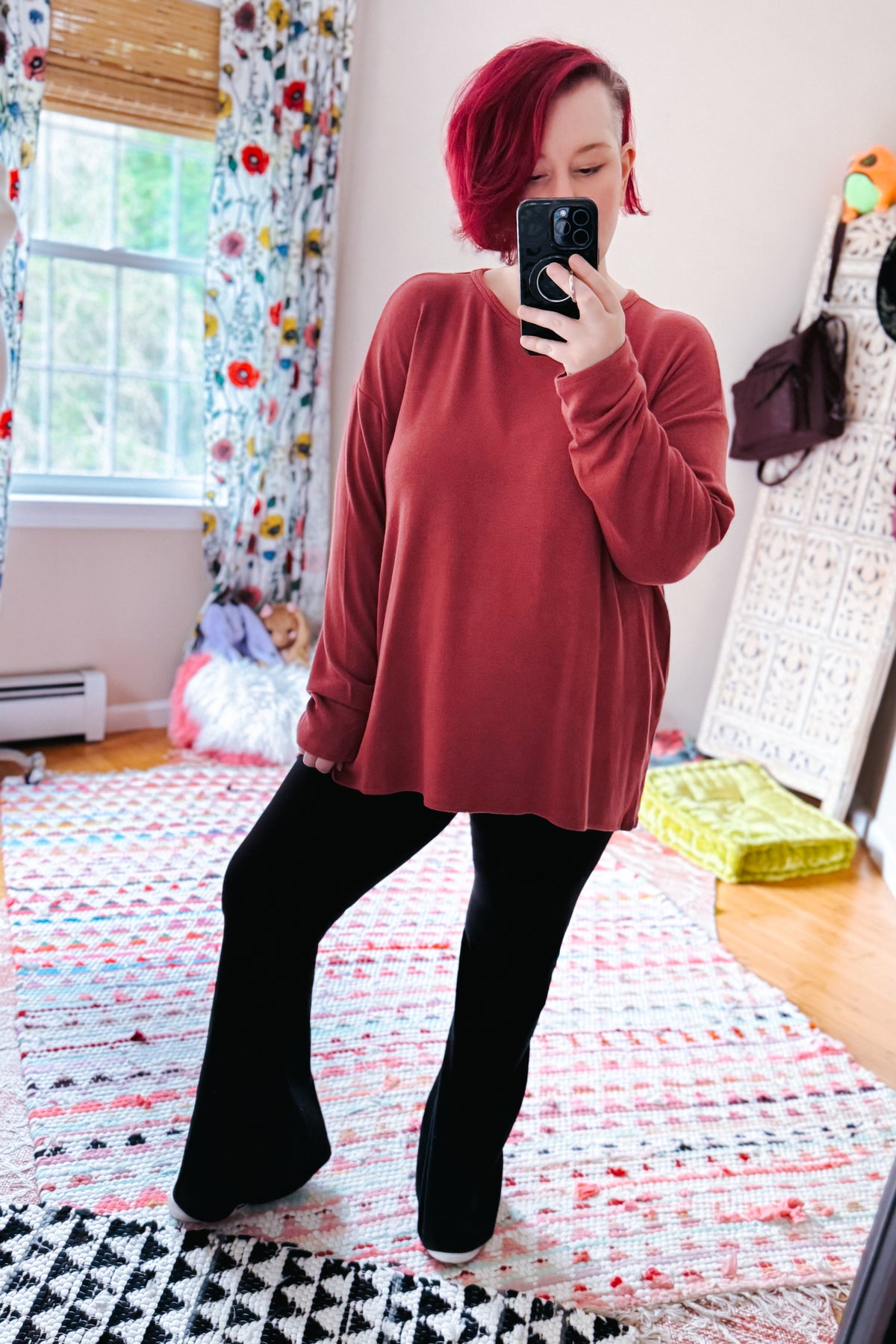 Woman wearing a cranberry long sleeve top with dropped shoulders and black pants in a colorful bedroom mirror selfie
