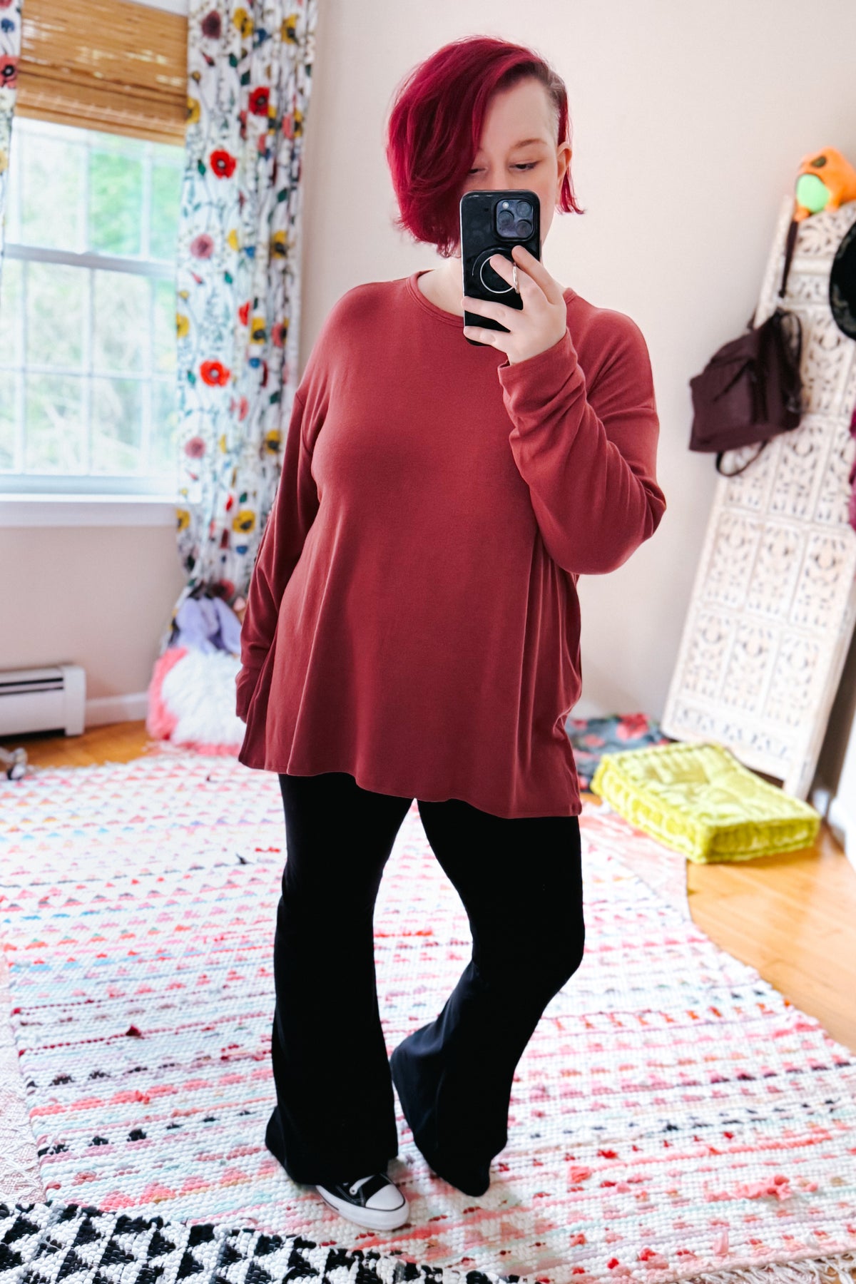 Woman in a cranberry long sleeve top with a relaxed drop shoulder fit styled with black pants and sneakers in a colorful bedroom mirror selfie