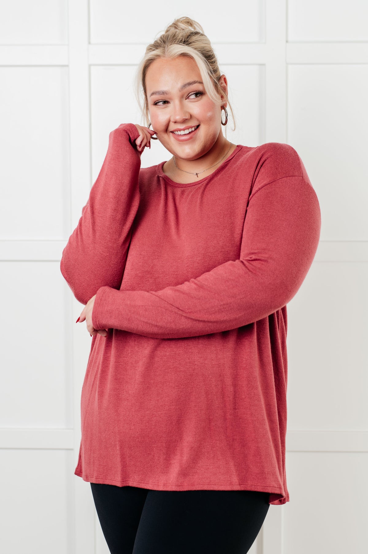 Smiling woman modeling a cranberry long sleeve top with a relaxed drop shoulder fit over black leggings indoors