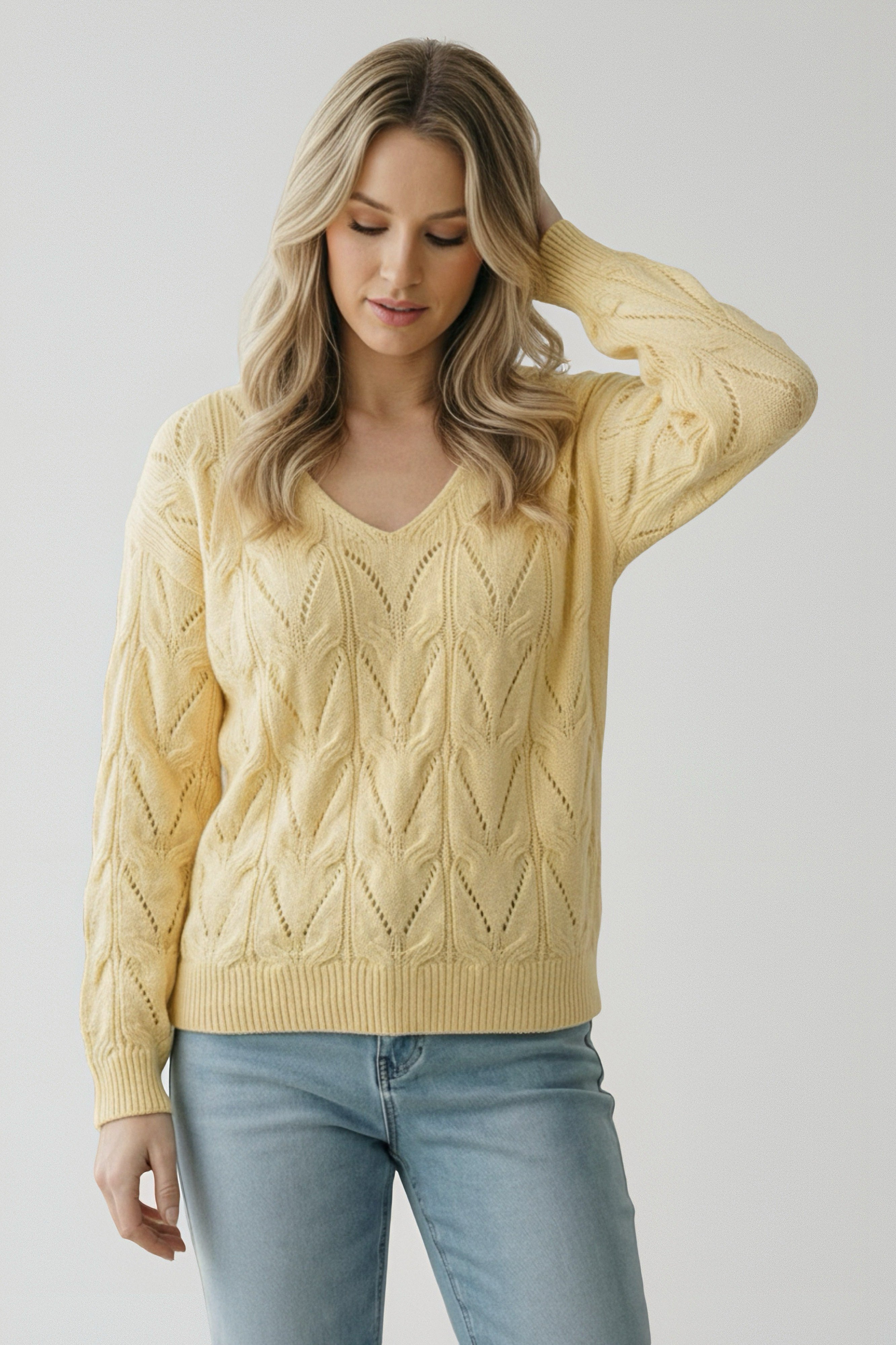 Woman wearing a soft yellow knit sweater with a delicate openwork pattern and light blue jeans, posing with one hand in hair