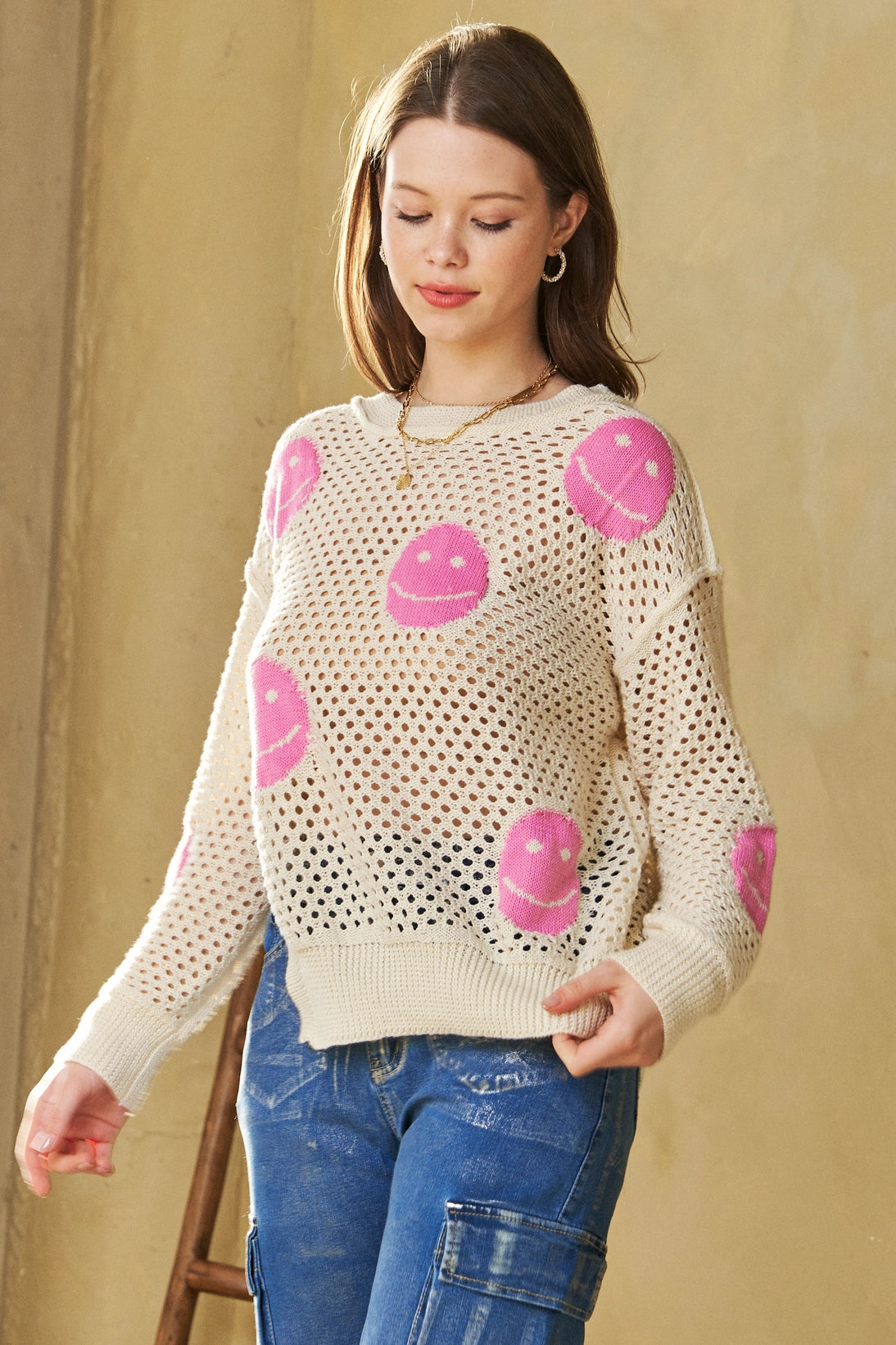 Woman wearing beige and pink In My Feelings Smiley Open Knit Top with bright pink smiley patterns and blue cargo pants in warm indoor lighting
