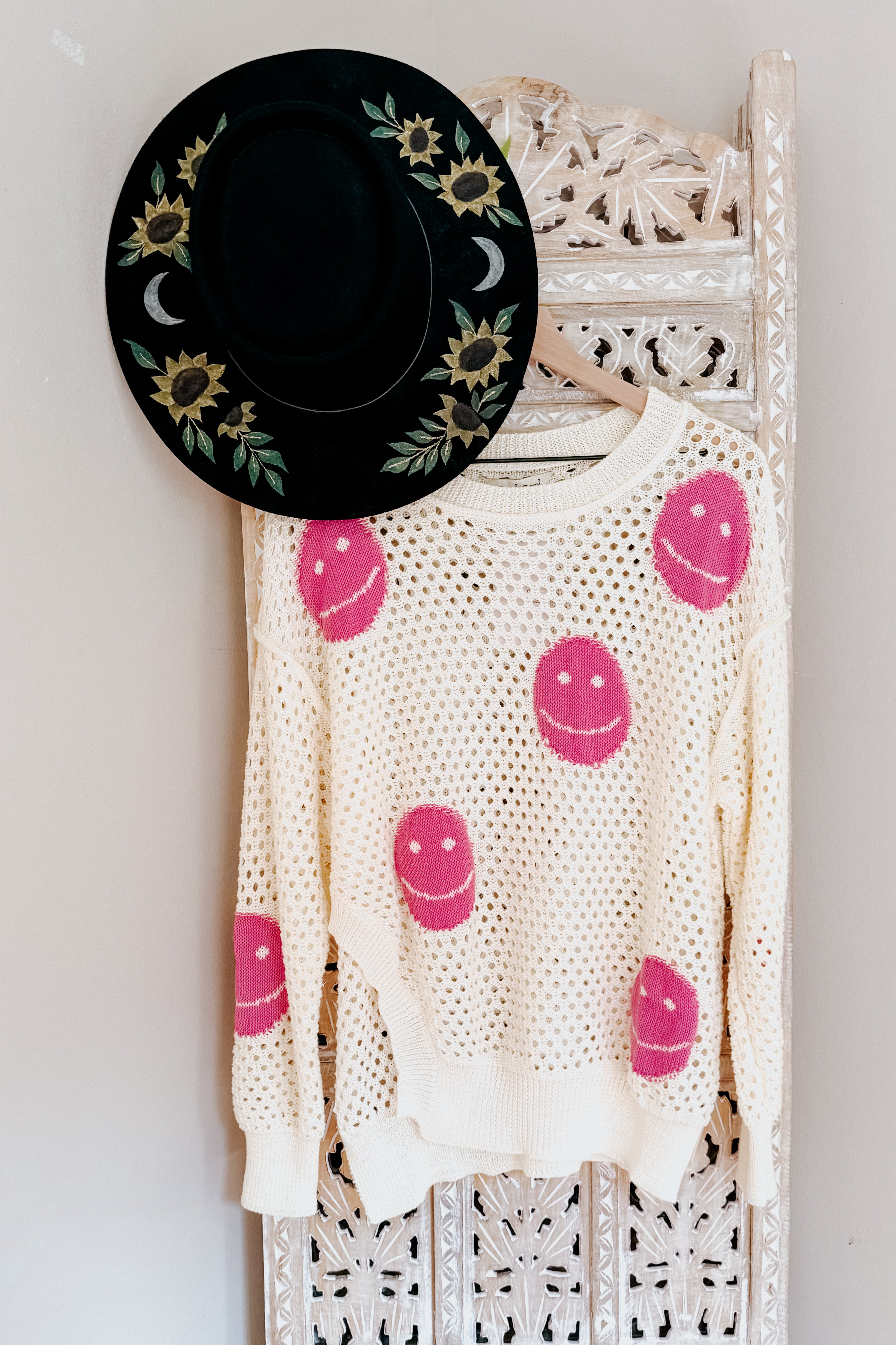 Beige and pink open knit top with smiley face patterns hanging on carved wooden panel with black floral wide-brim hat