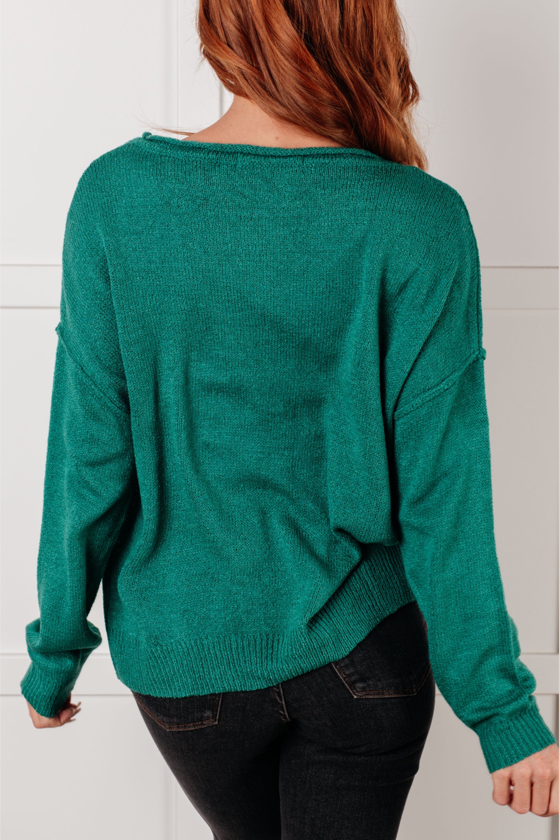 Woman seen from behind wearing a green In Stitches Drop Shoulder Sweater paired with dark jeans against a neutral wall background