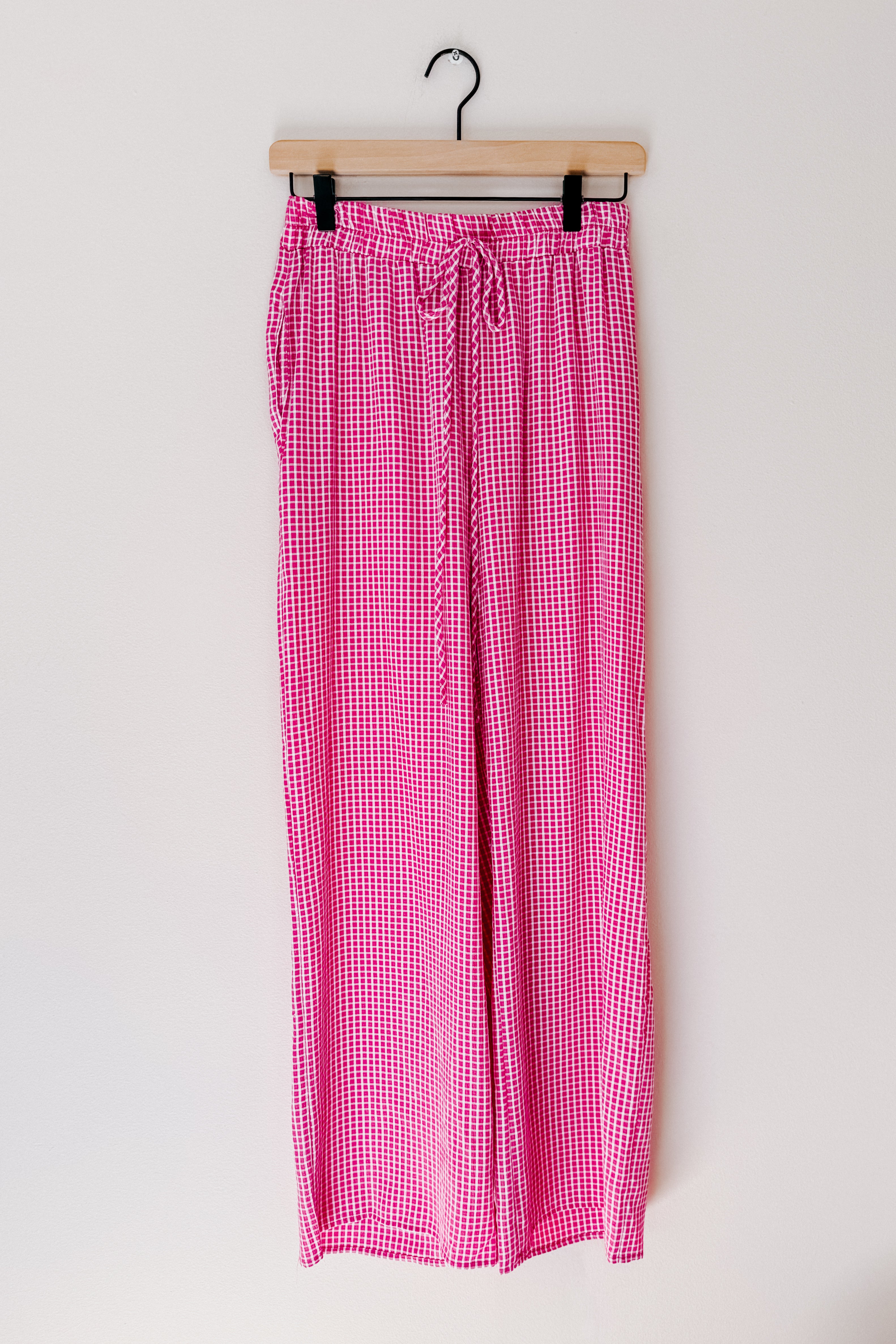 Pink checkered print pants with elastic waistband and drawstring hanging on wooden hanger against white background