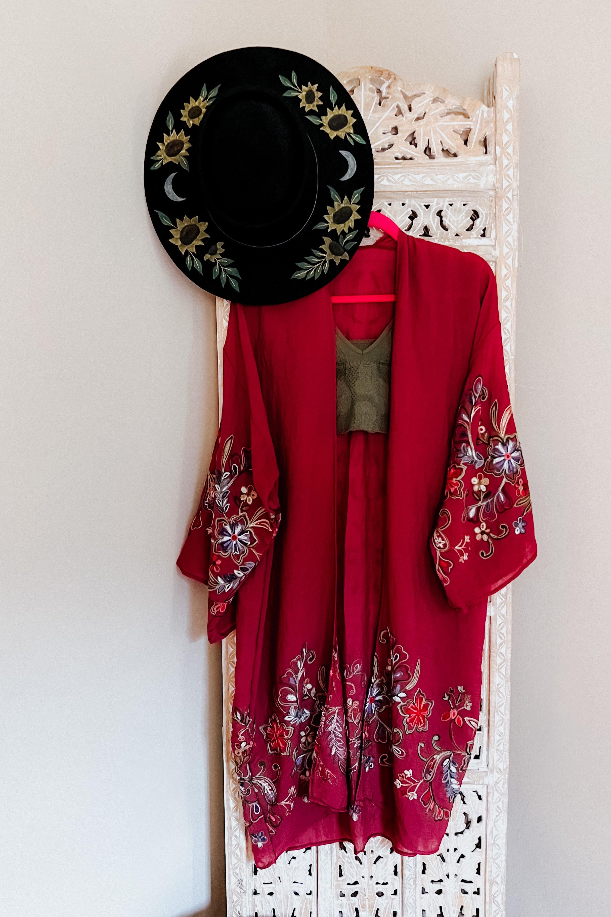 Crimson In The Meadow embroidered duster with floral designs on sleeves and hem hanging on decorative white screen with black hat