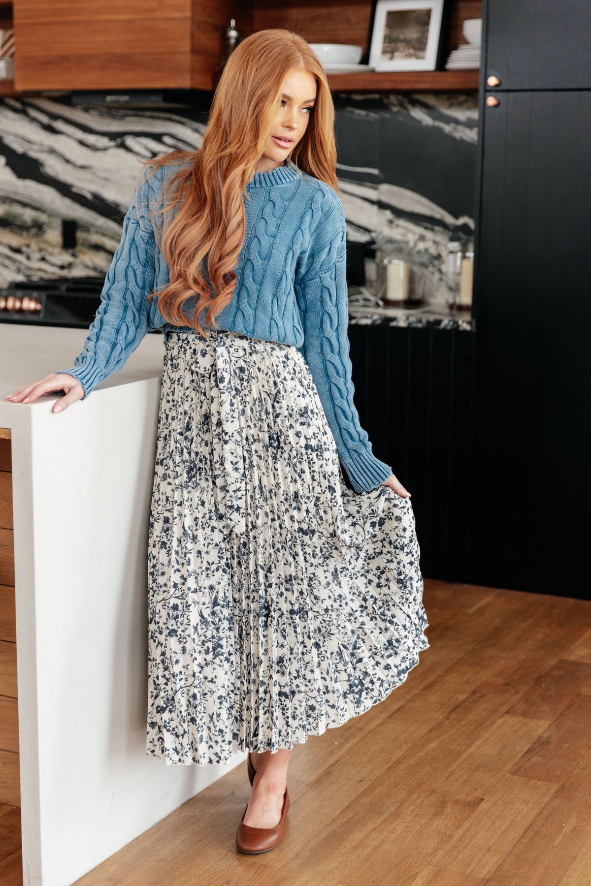 Woman wearing a blue mineral wash cable knit sweater with a patterned midi skirt and brown shoes in a modern kitchen