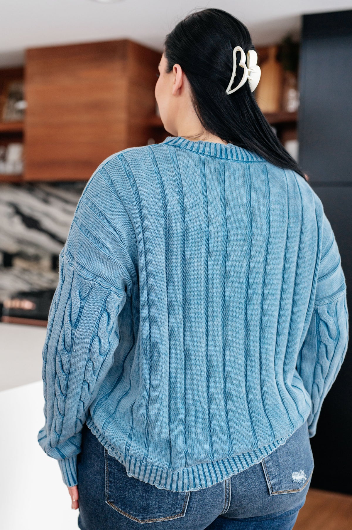 Woman seen from behind wearing a blue mineral wash cable knit sweater with textured sleeves jeans and a white hair clip indoors