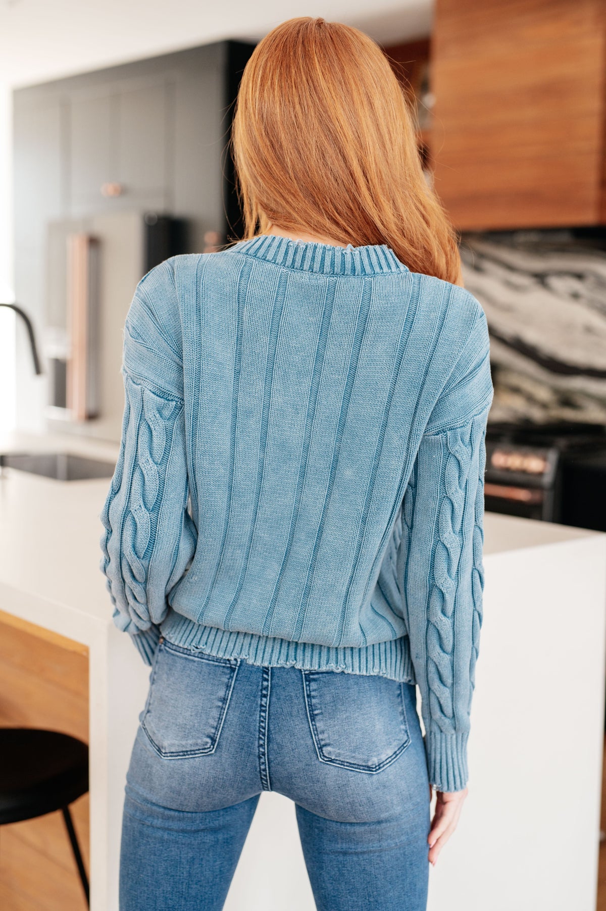 Woman shown from behind wearing a blue mineral wash cable knit sweater with textured sleeves and jeans in a modern kitchen
