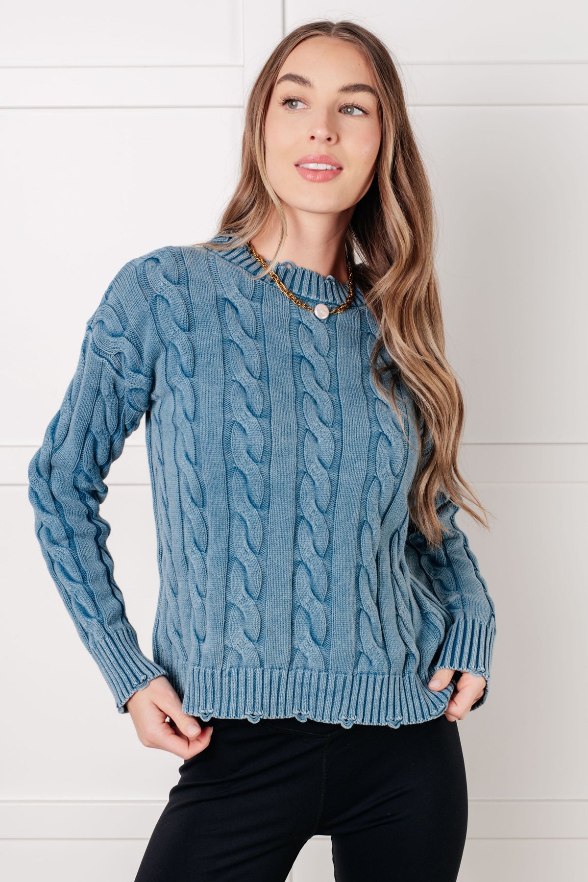 Model wearing a blue mineral wash cable knit sweater with distressed ribbed trim and long sleeves paired with black pants indoors