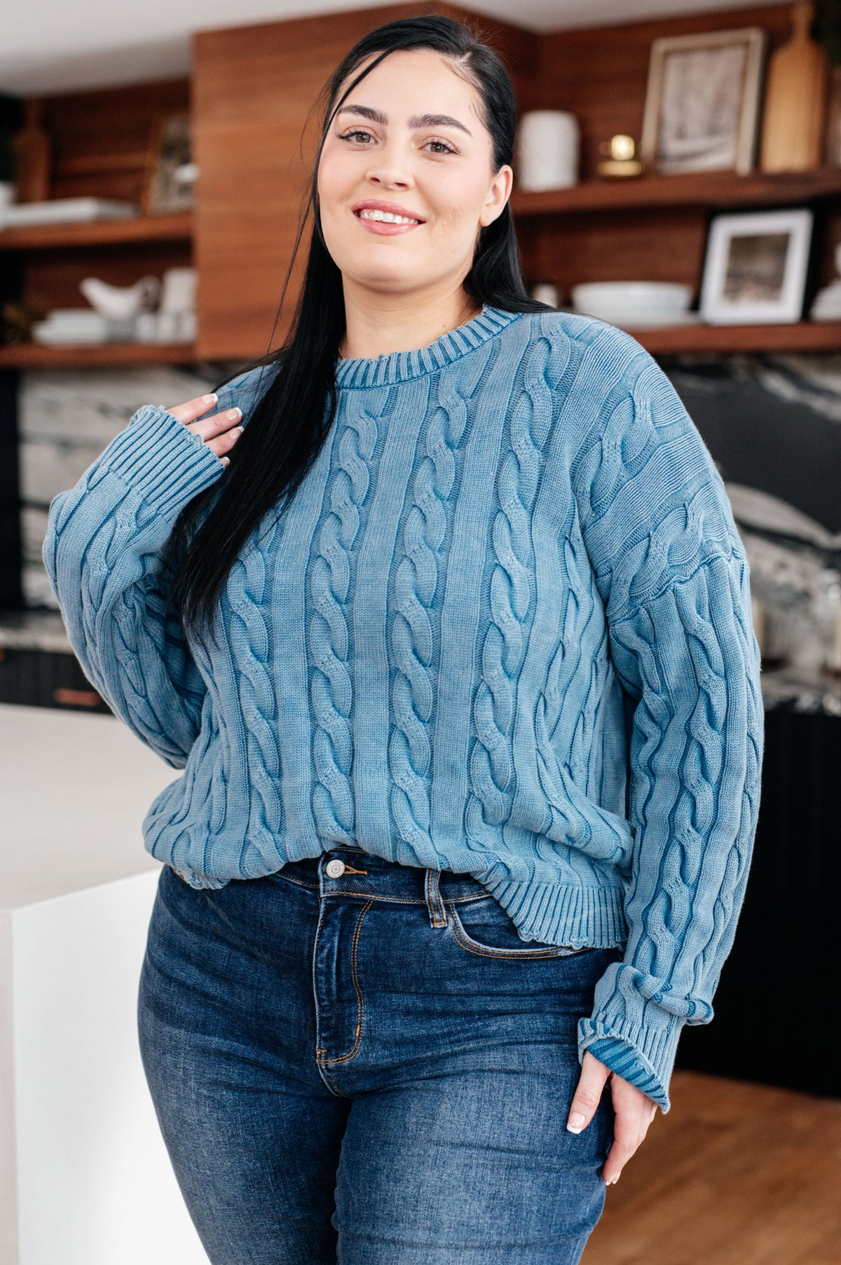 Woman wearing a blue cable knit sweater with a ribbed crew neckline and cuffs paired with jeans in a cozy home interior