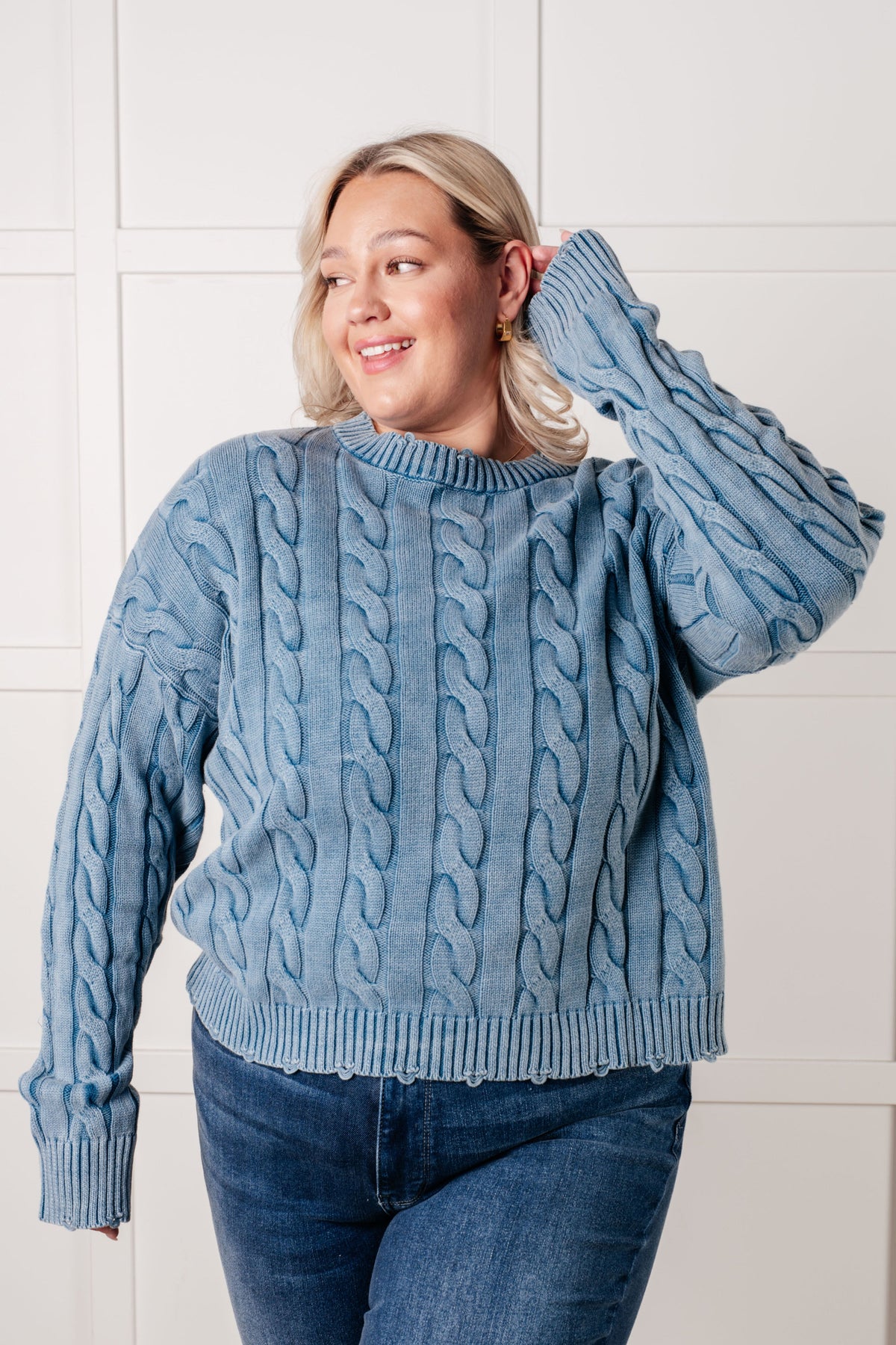 Smiling woman in a blue cable knit sweater with ribbed trim touches her hair while posing in jeans against a paneled wall
