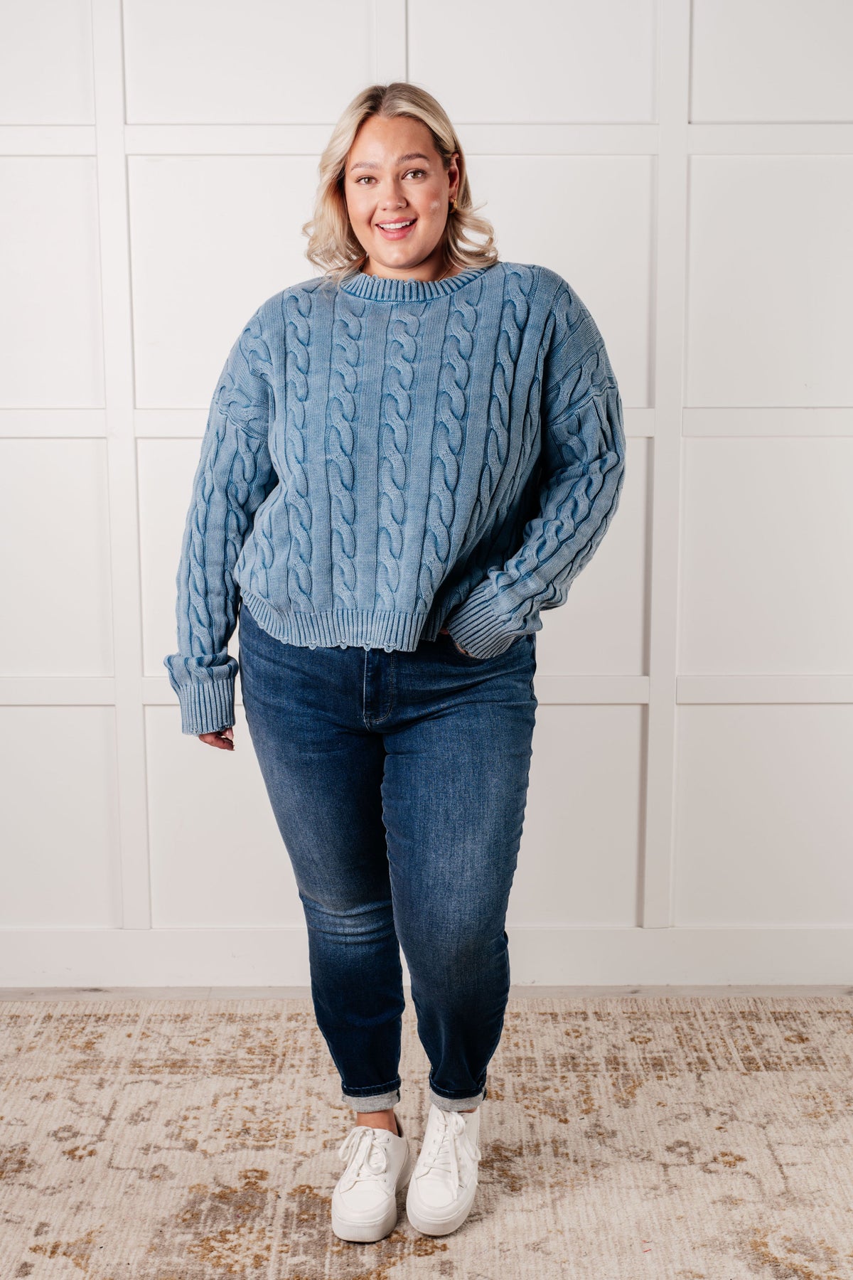 Woman wearing a blue mineral wash cable knit sweater with distressed ribbed trim styled with jeans and white sneakers indoors