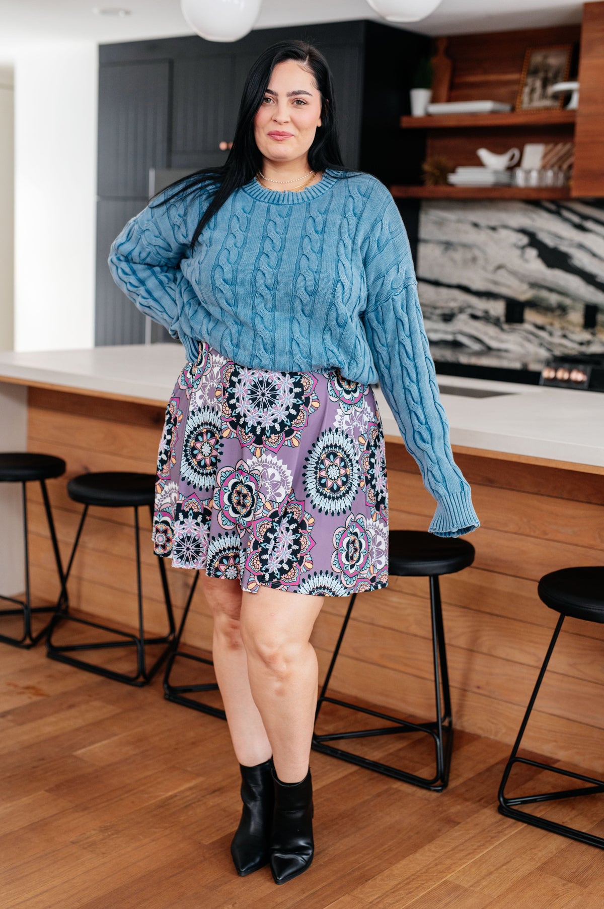 Woman wearing a blue cable knit sweater with relaxed sleeves and a patterned skirt with black ankle boots in a modern kitchen