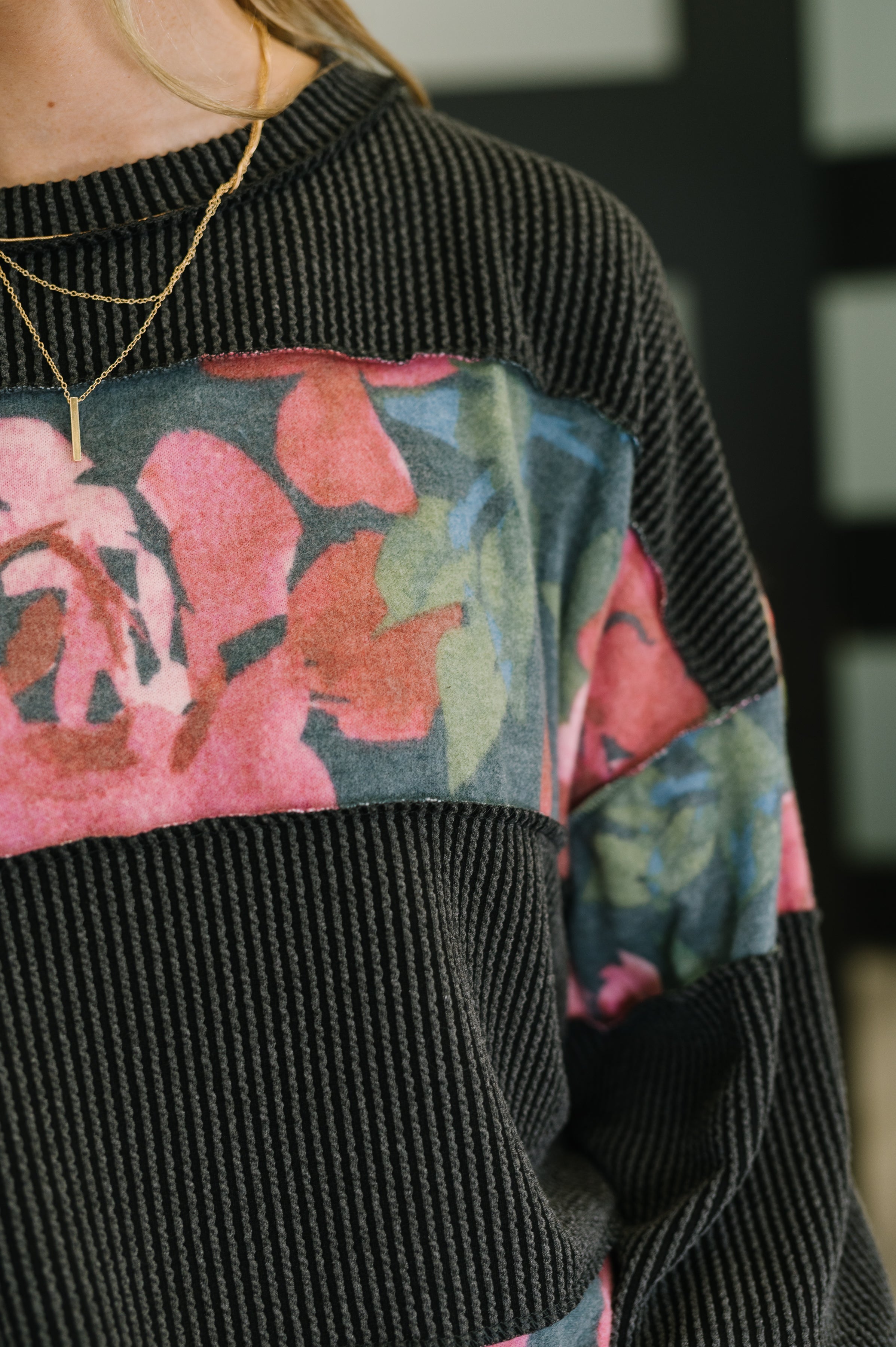 Close-up of black textured pullover with wide floral print panel across chest and sleeves worn with layered gold necklaces